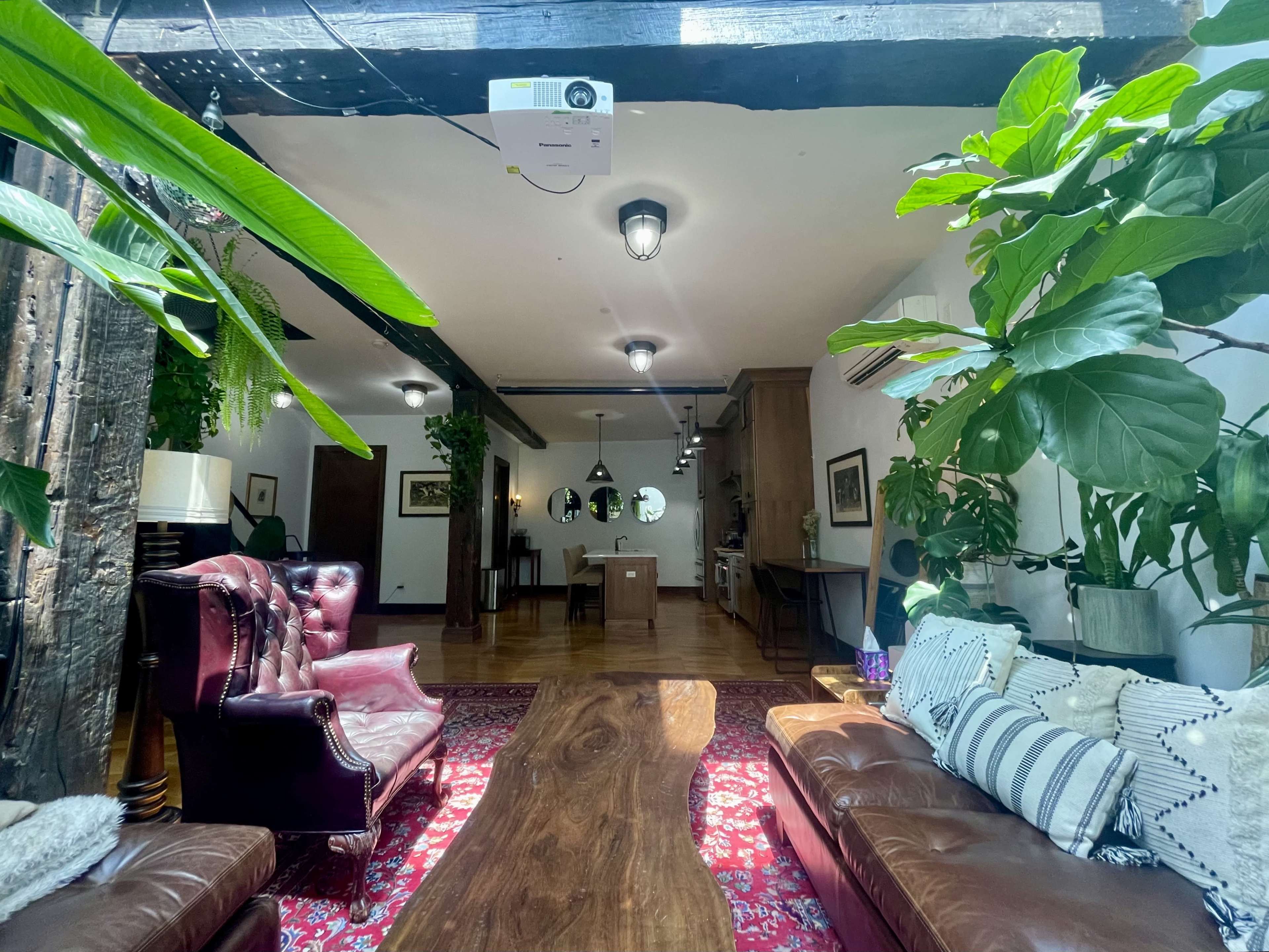 A living space featuring two brown leather sofas, a large wooden coffee table, and numerous plants, with an open view towards a dining area and kitchen.