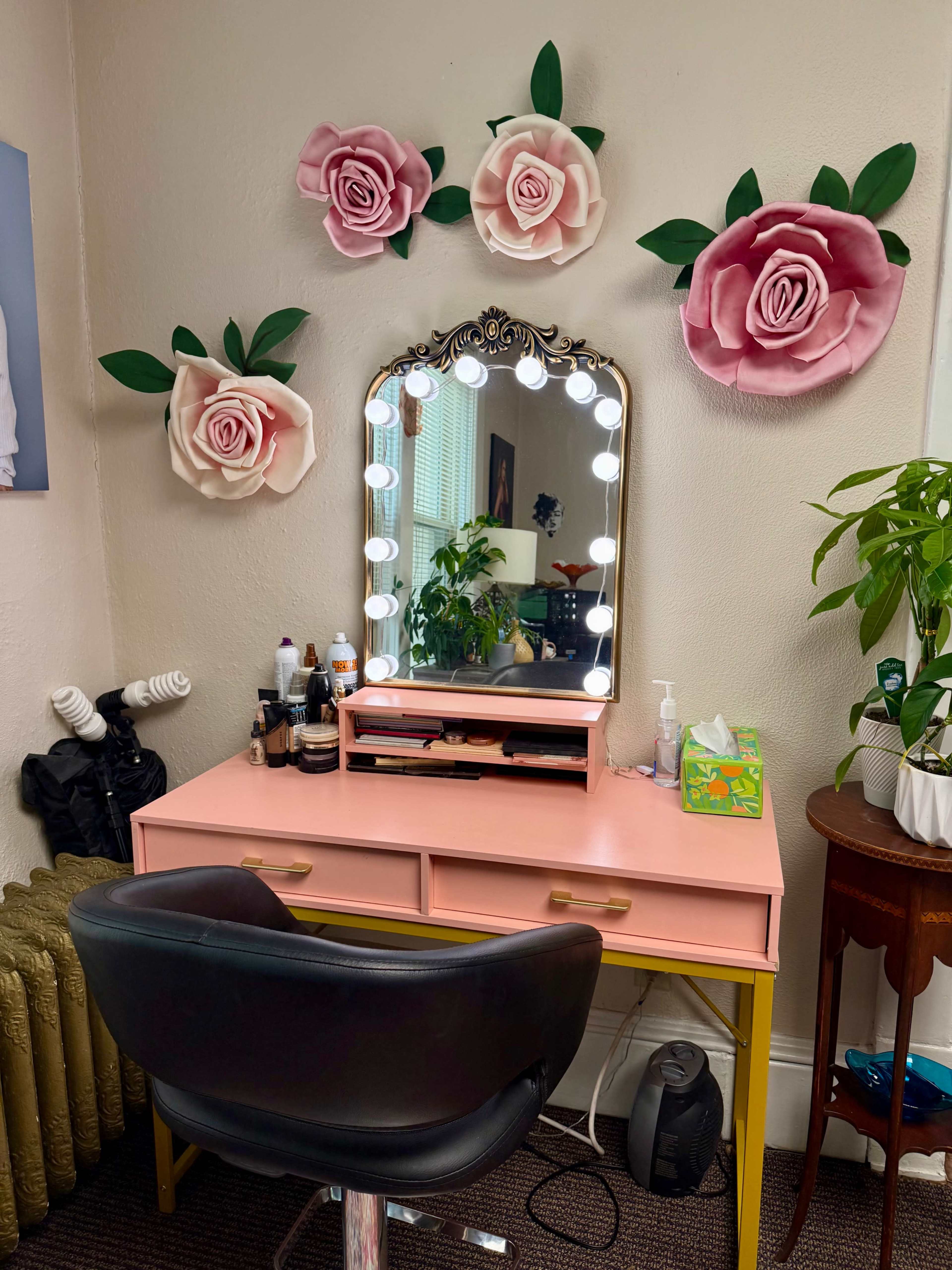 The image shows a vanity setup with a large mirror surrounded by lights, a pink desk with drawers, and decorative paper flowers on the wall.