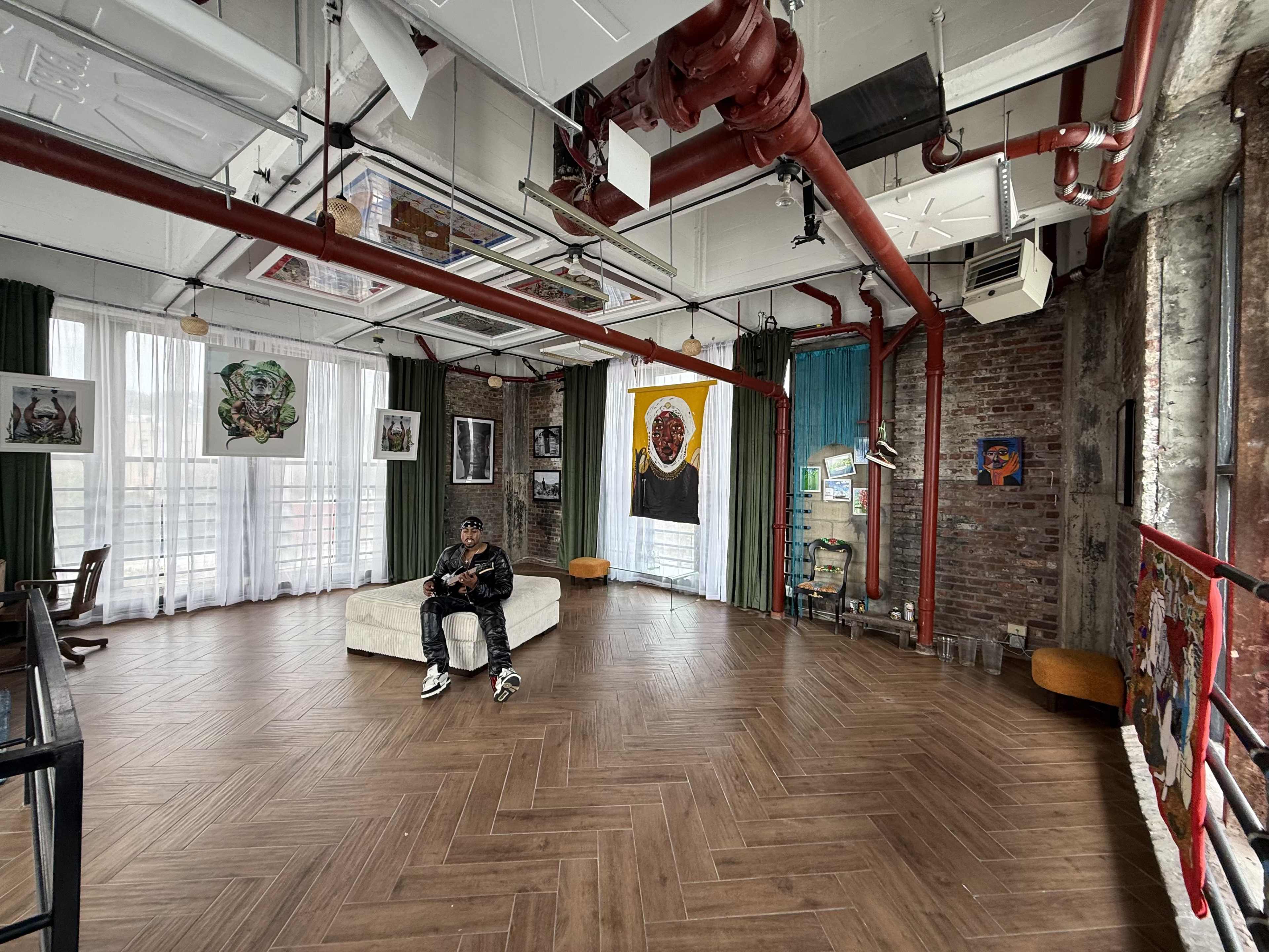 The image depicts a spacious, industrial-style room with hardwood floors, large windows, and eclectic artwork on the walls, featuring a person sitting on a white couch.