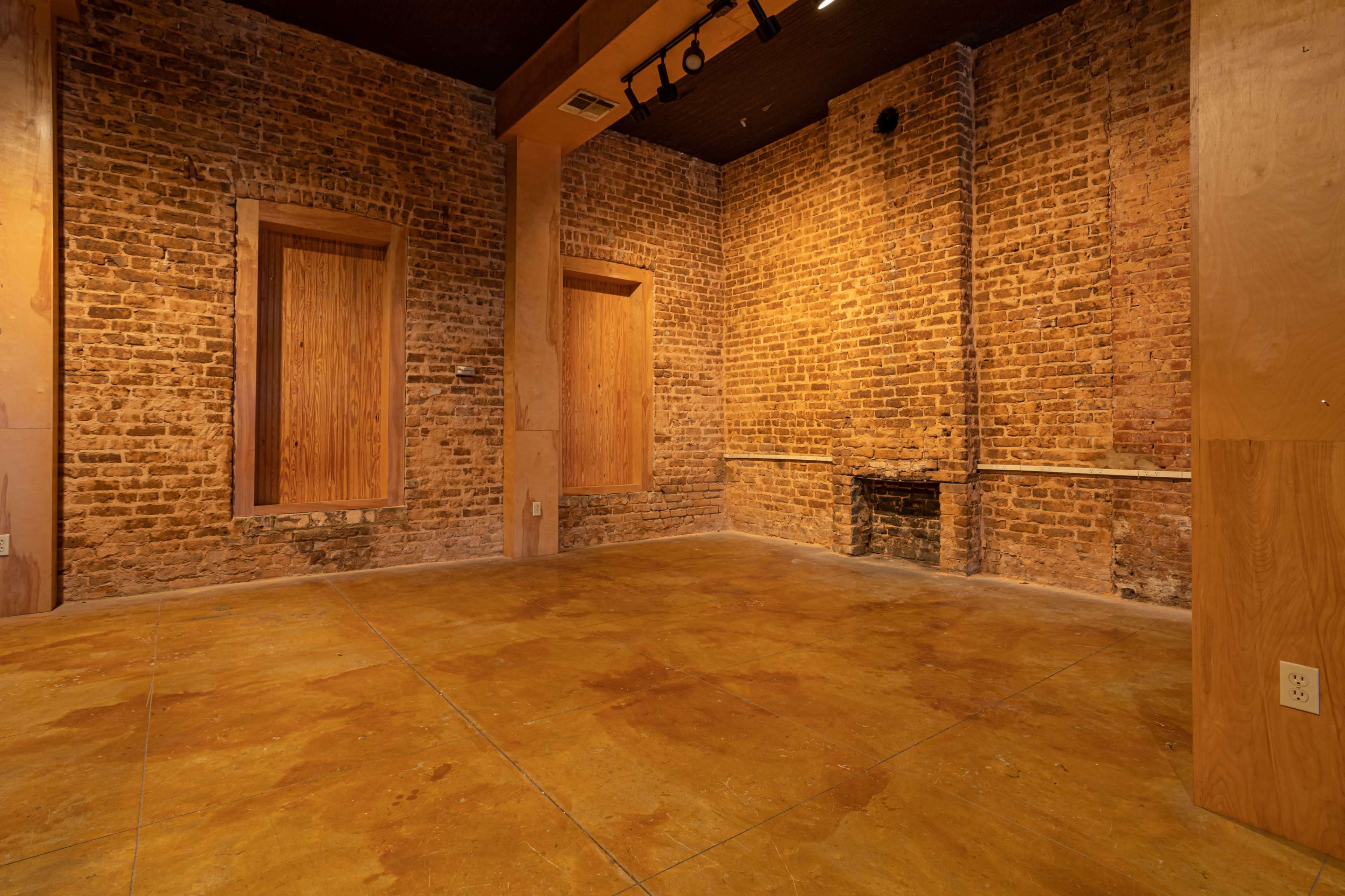 The image shows a vacant room with exposed brick walls, wooden paneling over windows, and a concrete floor.