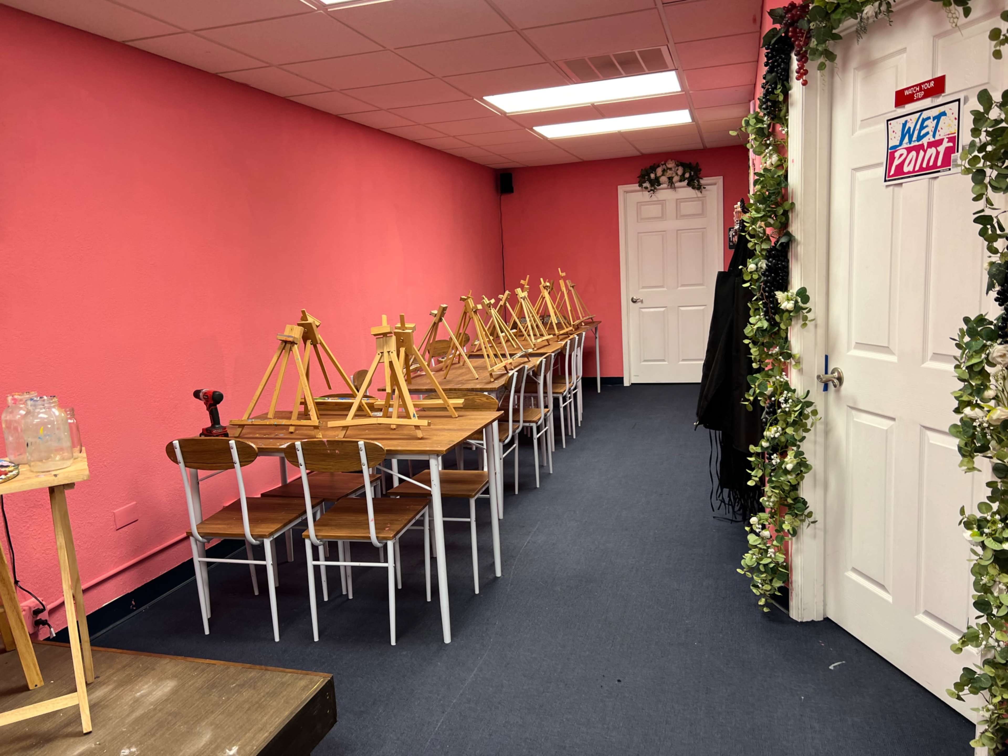 A classroom setting with several wooden easels arranged on tables against a pink wall, leading to a door labeled "Wet Paint."