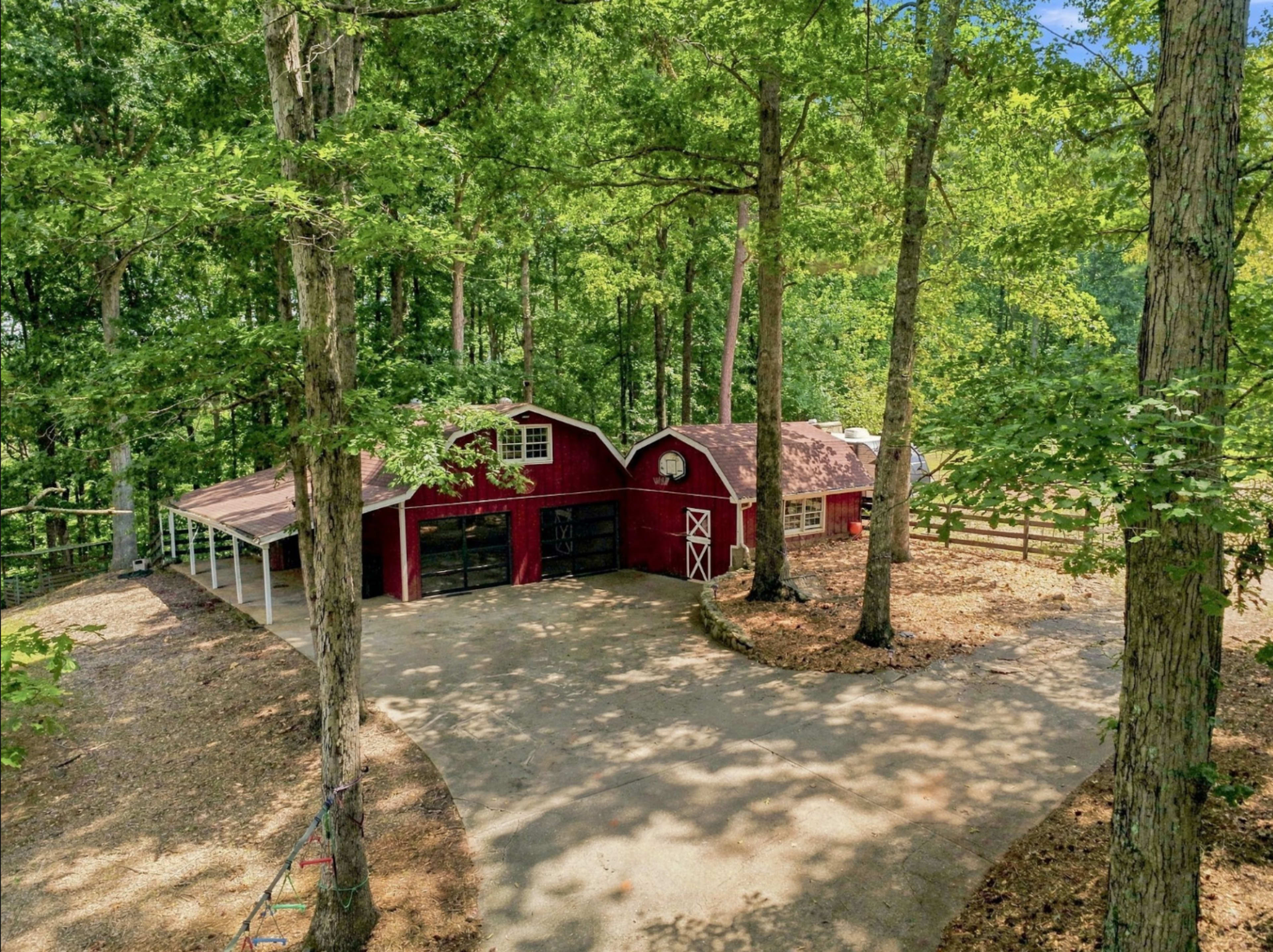 A red barn with a circular driveway is surrounded by tall trees in a wooded area.