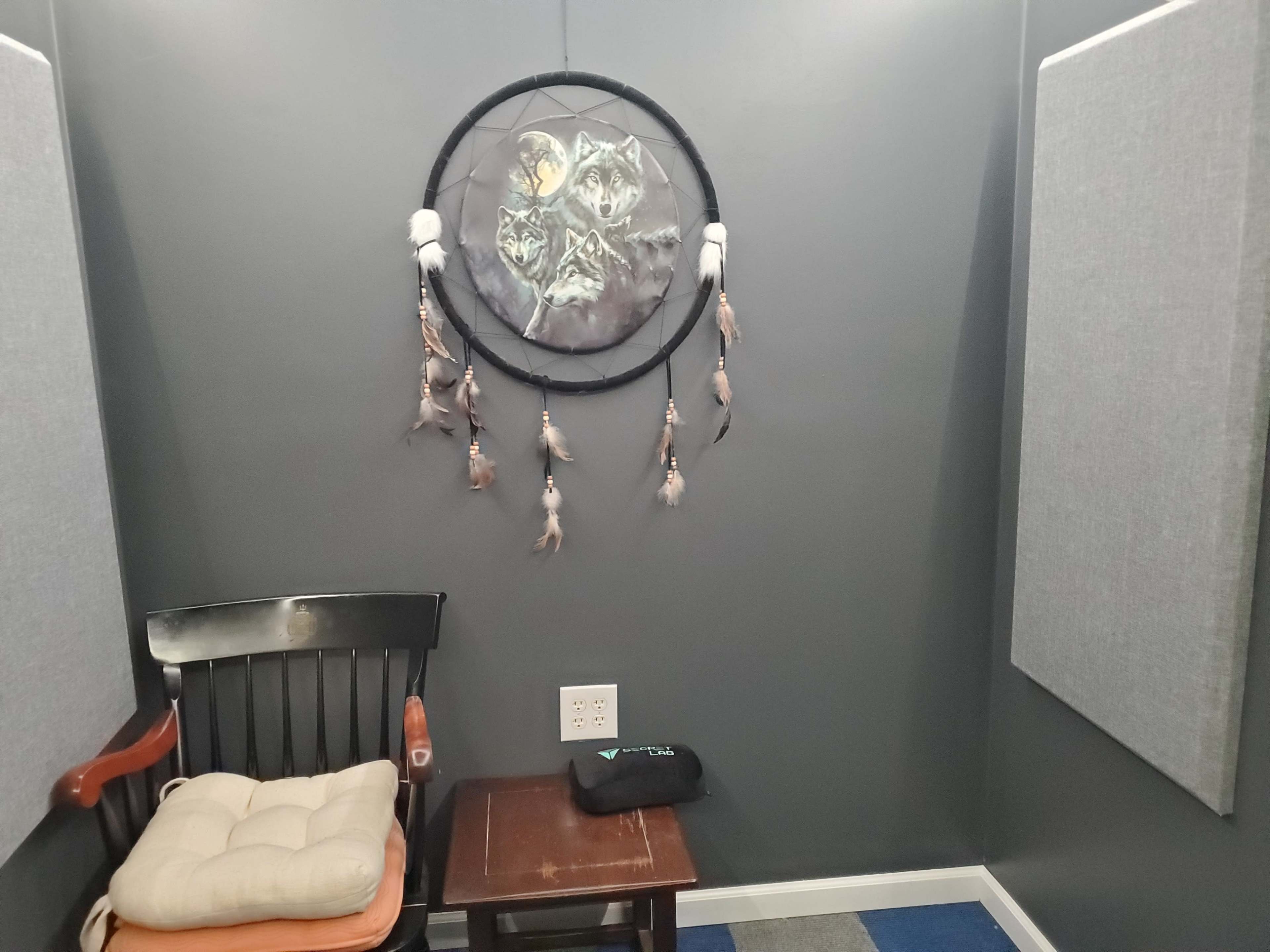A small room with gray walls, featuring a dreamcatcher with owl designs, a wooden chair with a cushion, and a small table beside it.