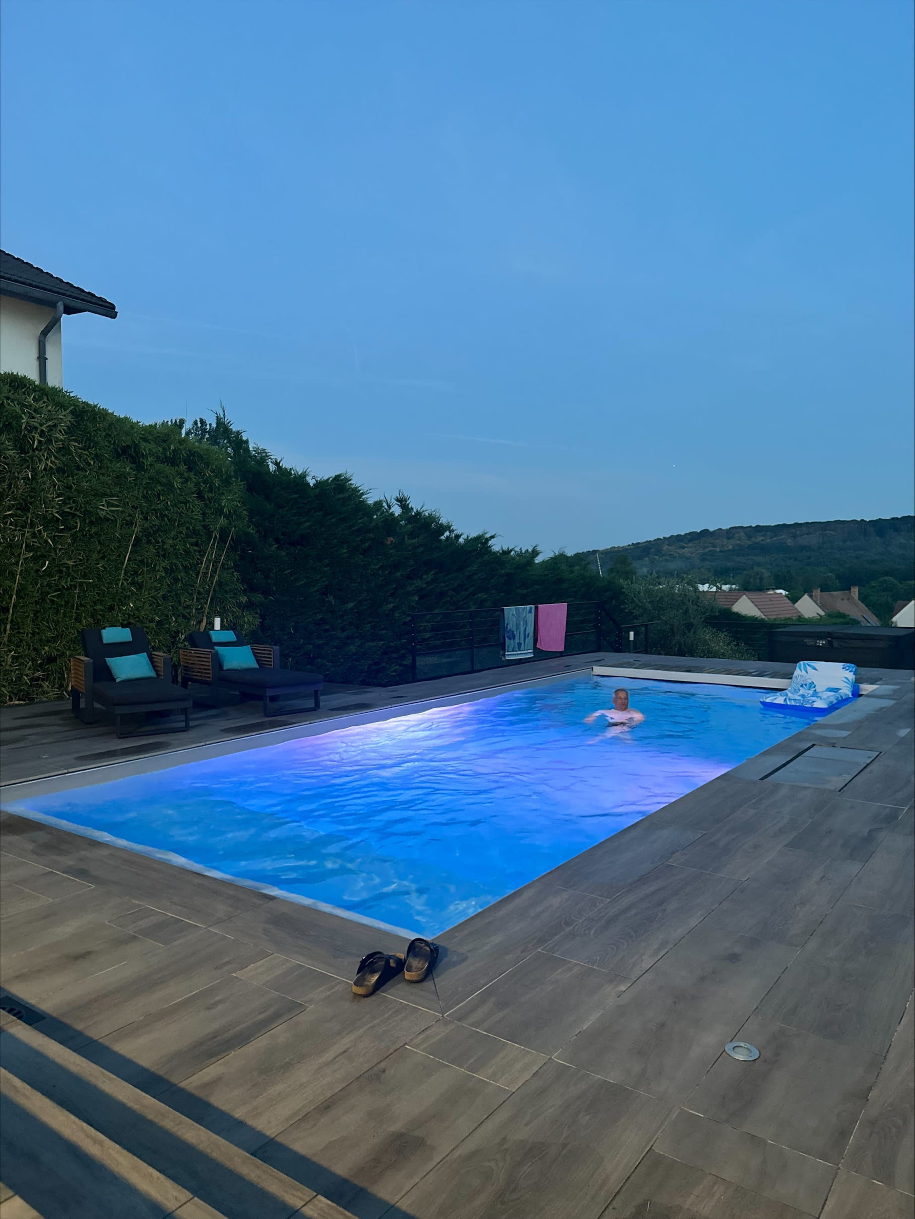 A person swims in a well-lit pool surrounded by greenery and lounge chairs at twilight.