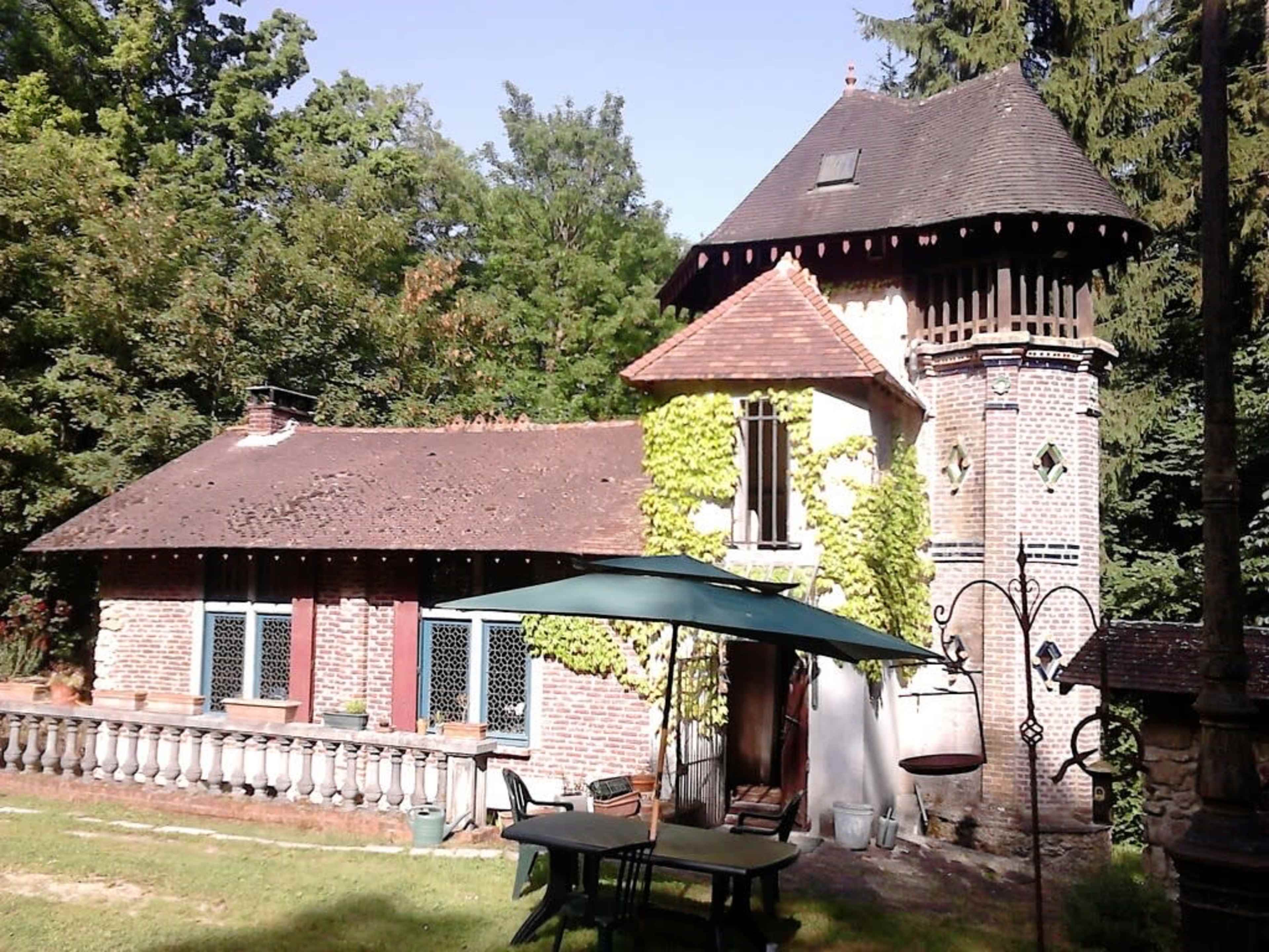 A brick house with a tower is surrounded by trees and features a green lawn and outdoor furniture.