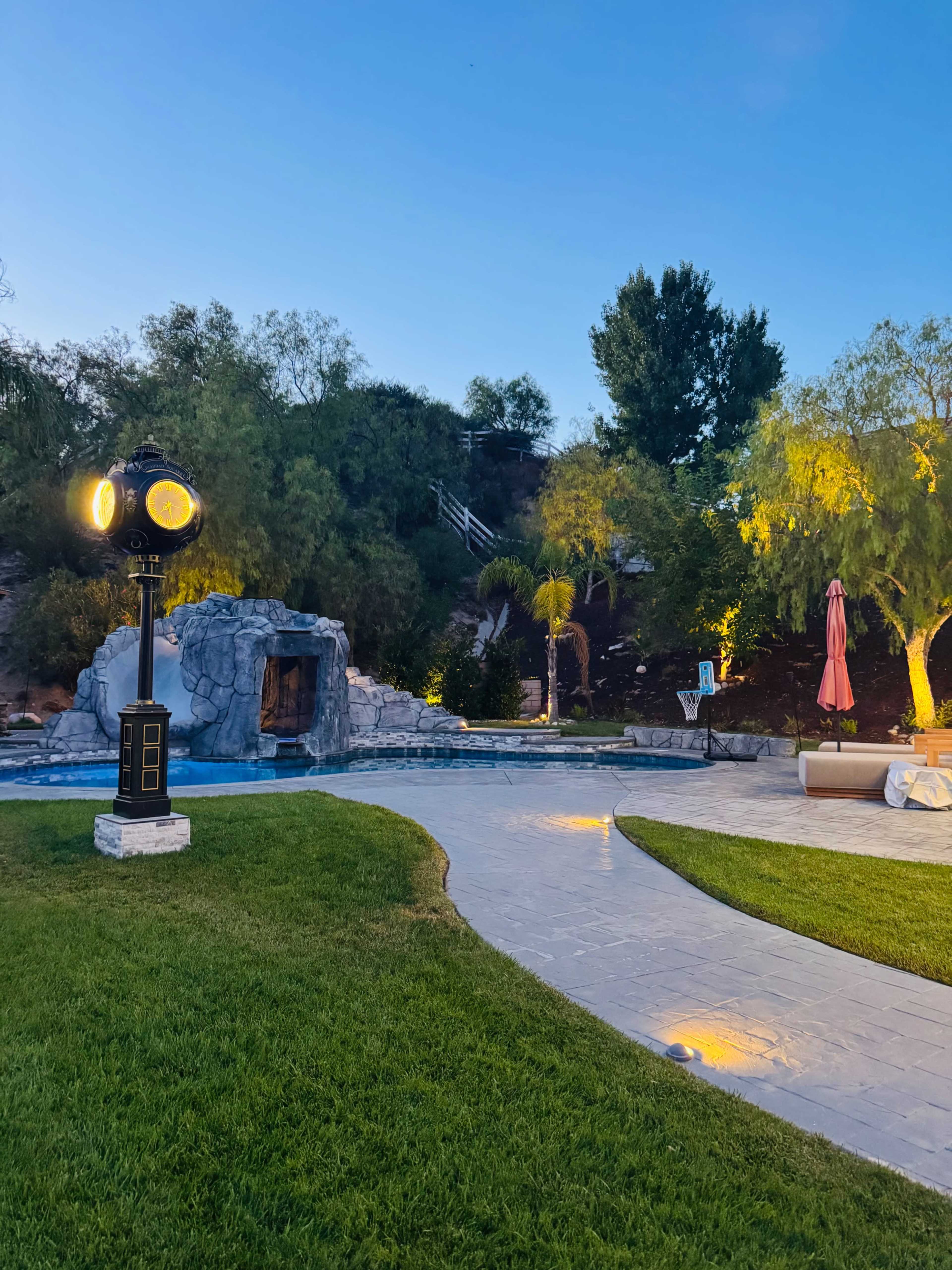 The image shows a well-landscaped backyard featuring a swimming pool with a rock waterfall, decorative lighting, and a path leading through a grassy area.