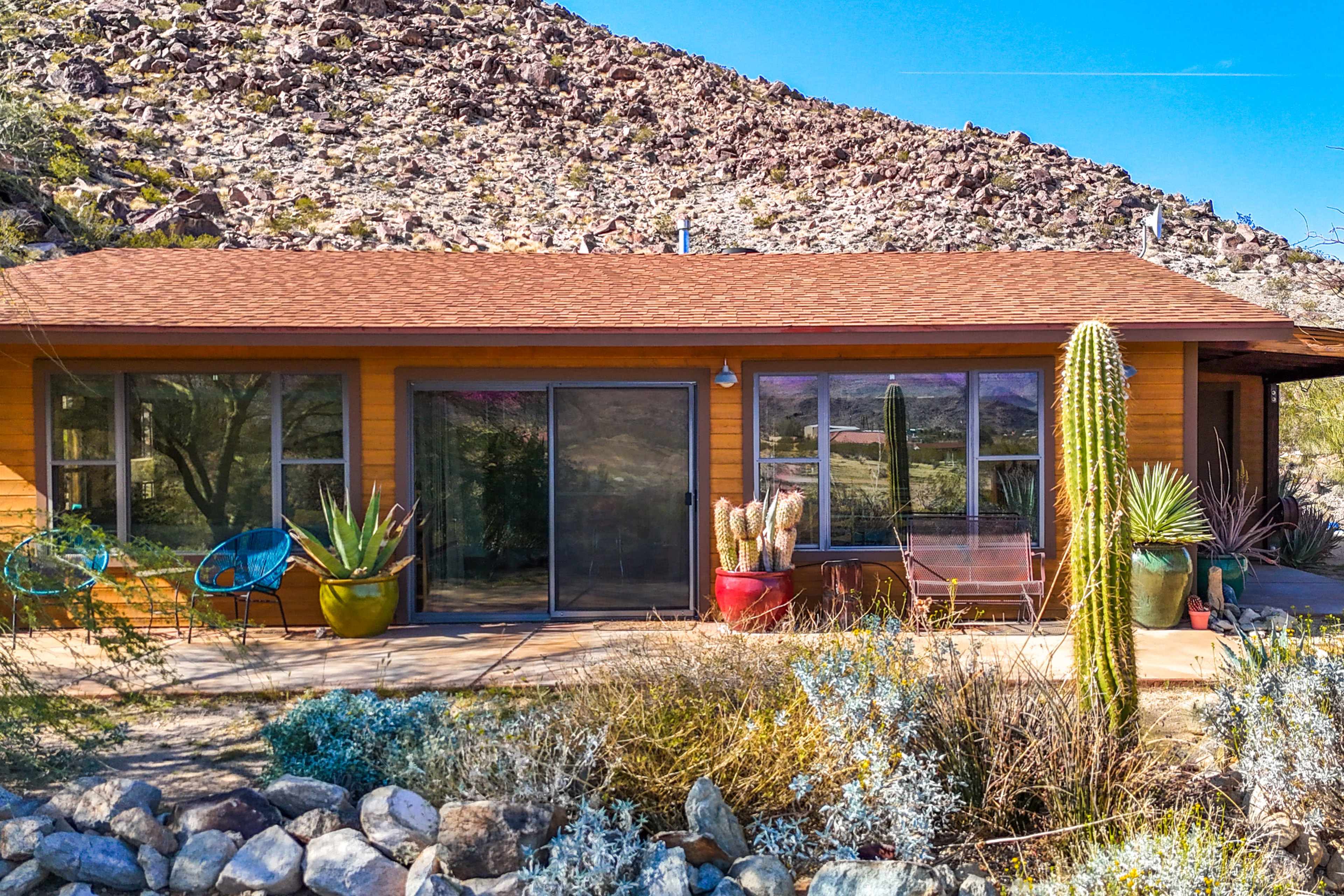 A single-story wooden cabin with large glass sliding doors and colorful potted plants sits against a rocky hillside.