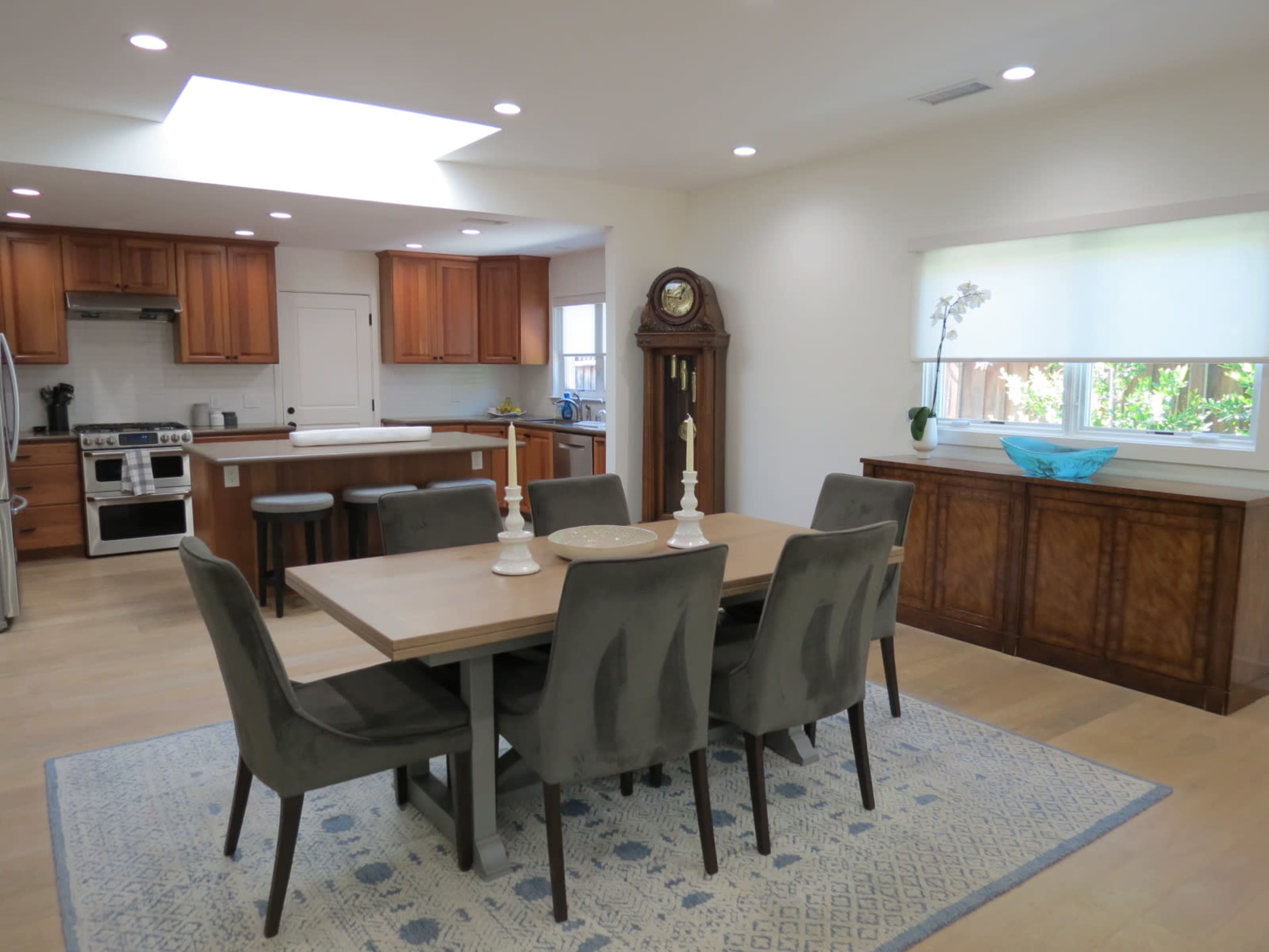 A modern dining area features a wooden table with gray chairs, surrounded by a well-lit kitchen with wooden cabinets and a clock against the wall.