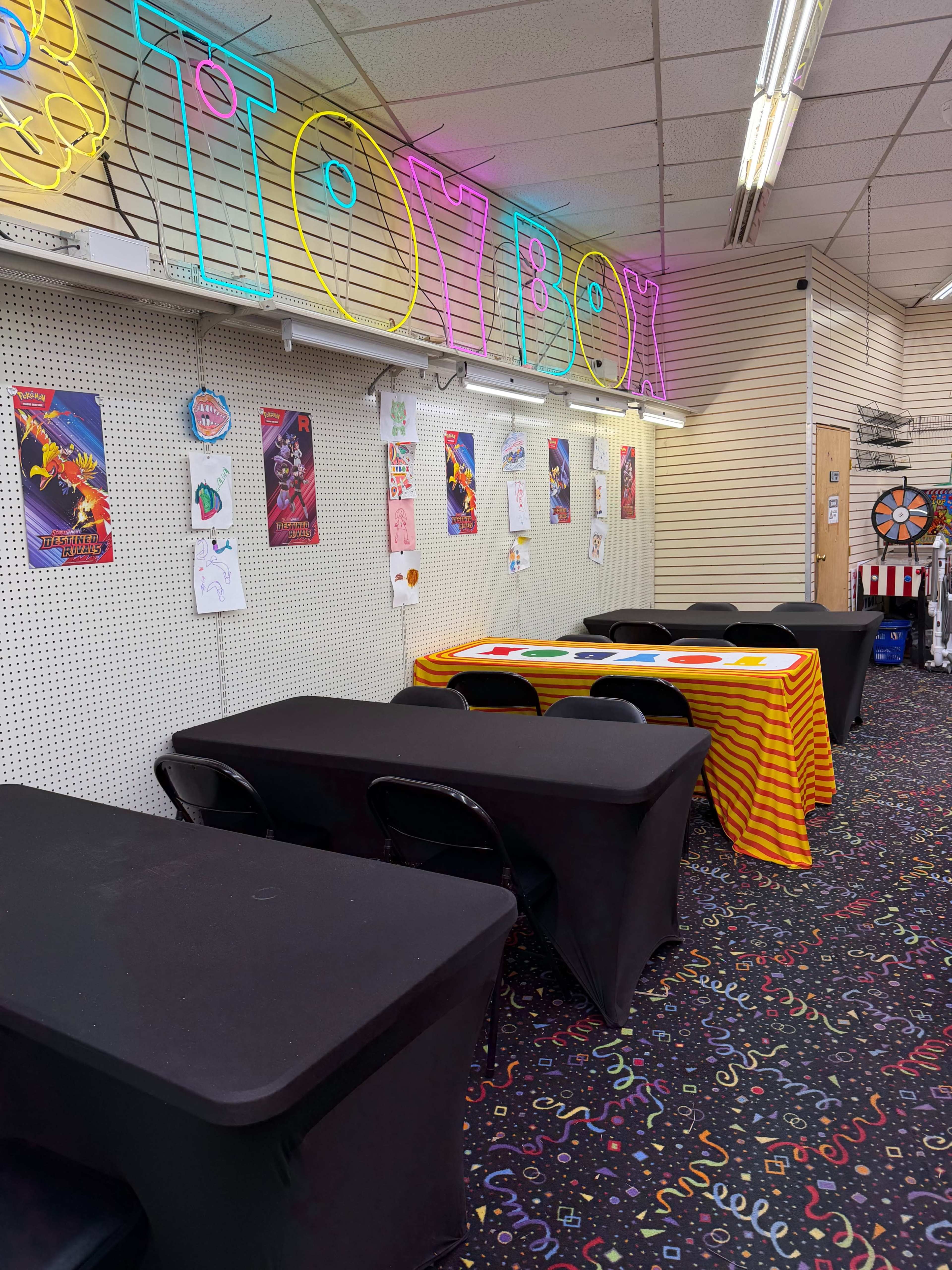 The image shows a brightly colored room with a neon "TOYBOX" sign, several tables covered with black and orange tablecloths, and decorated walls featuring various posters.
