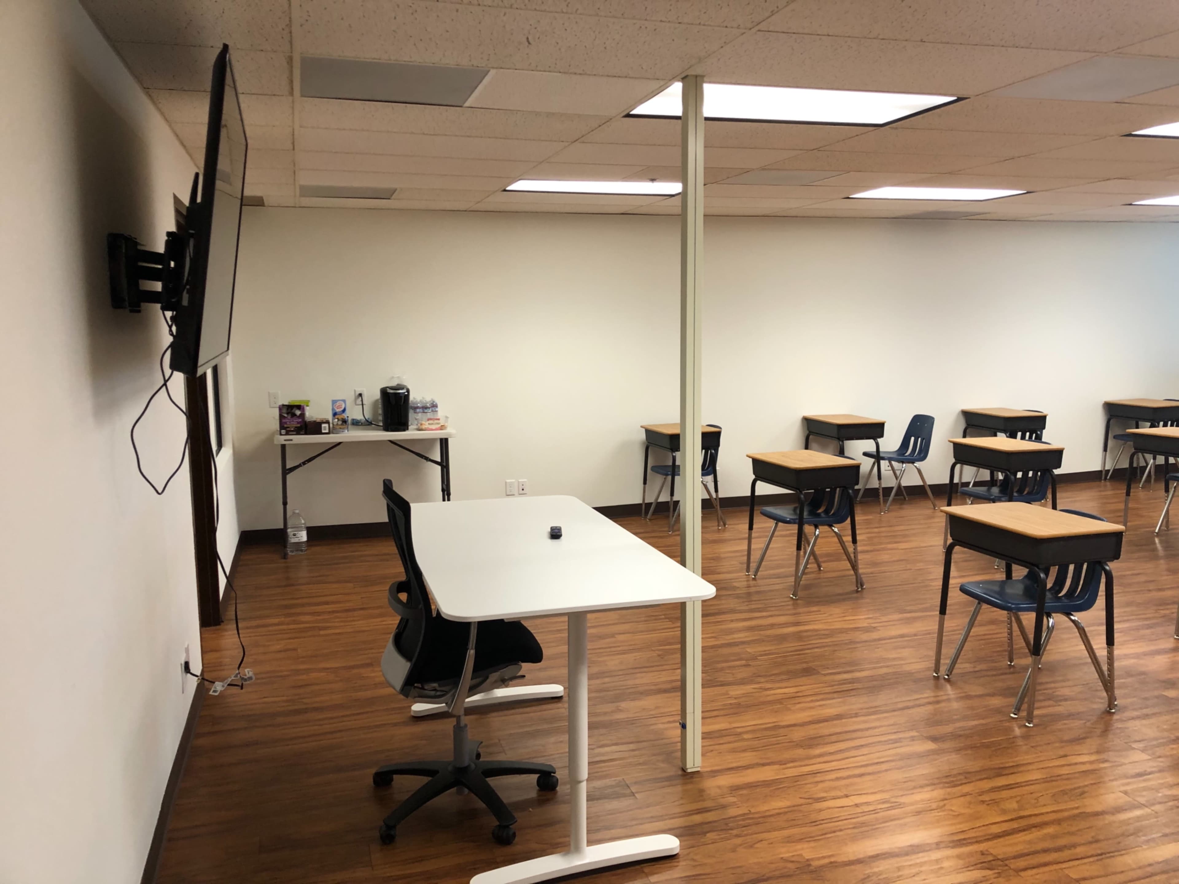 The image shows a classroom with several empty desks, a television mounted on the wall, and a table with a coffee maker in a corner.