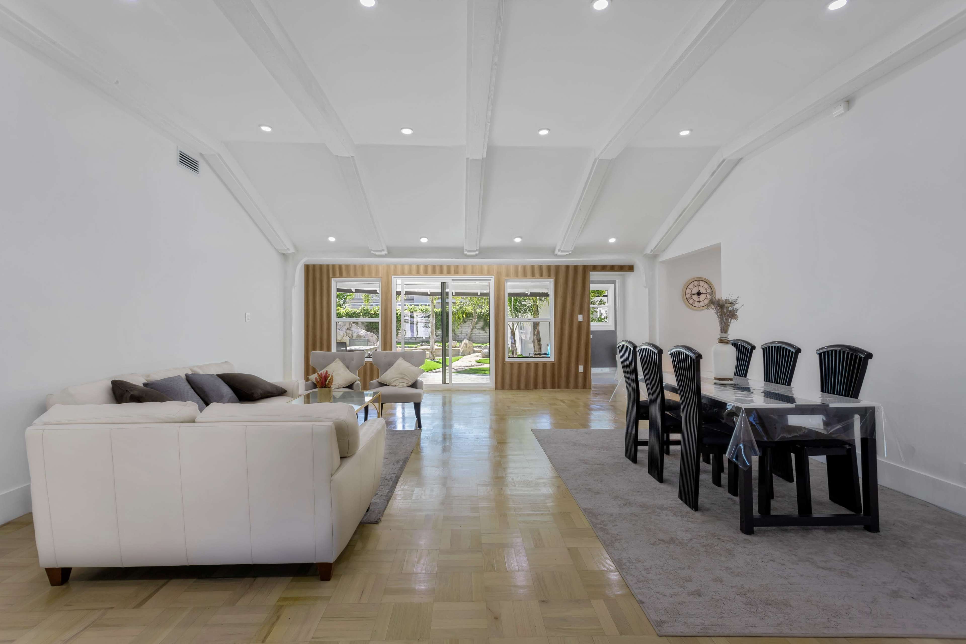 A spacious living area featuring a white sofa, a glass dining table with black chairs, and large windows that offer a view of an outdoor space.