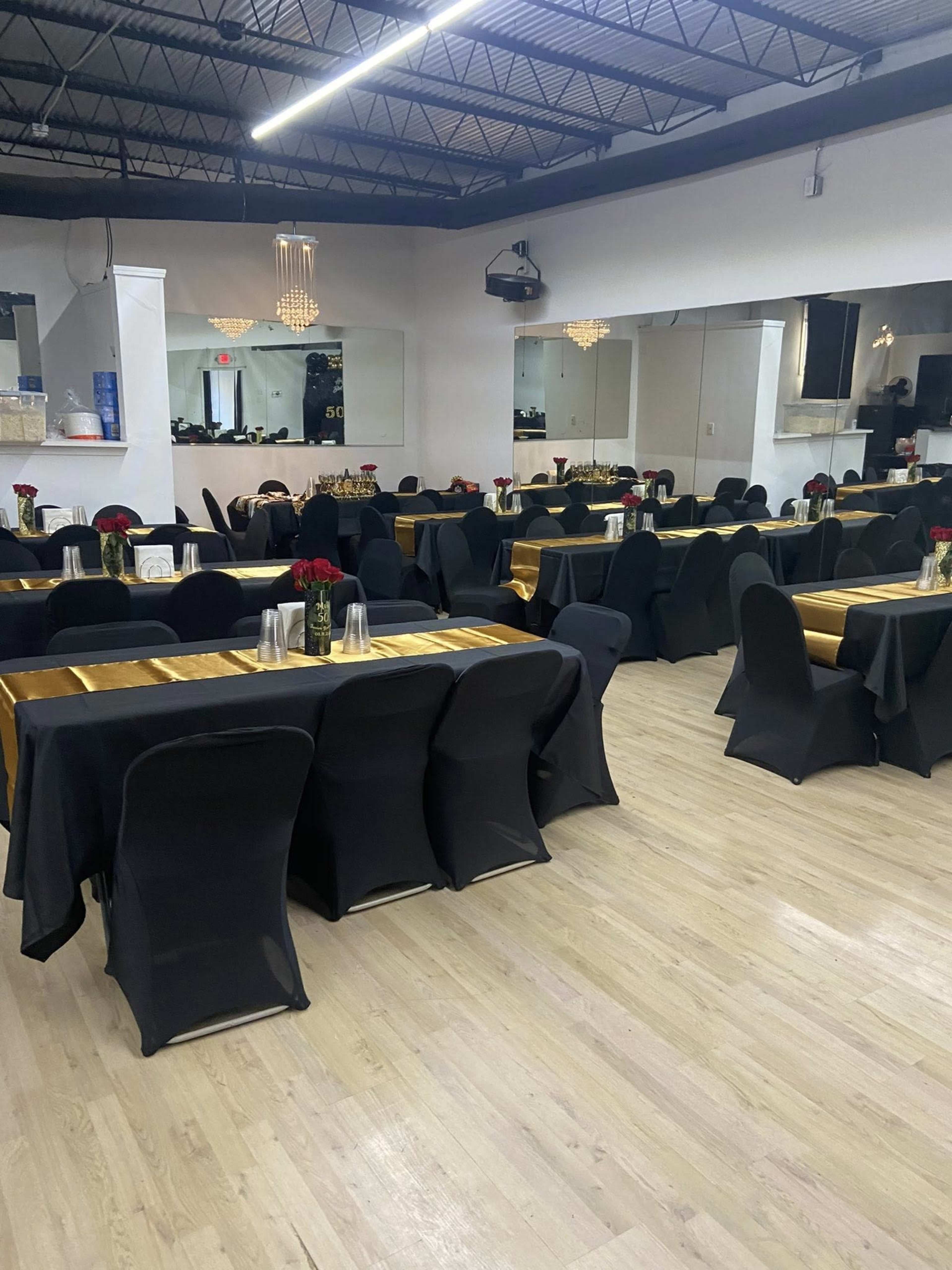 The image shows a neatly arranged dining area with several black-covered tables adorned with golden table runners and red rose centerpieces.