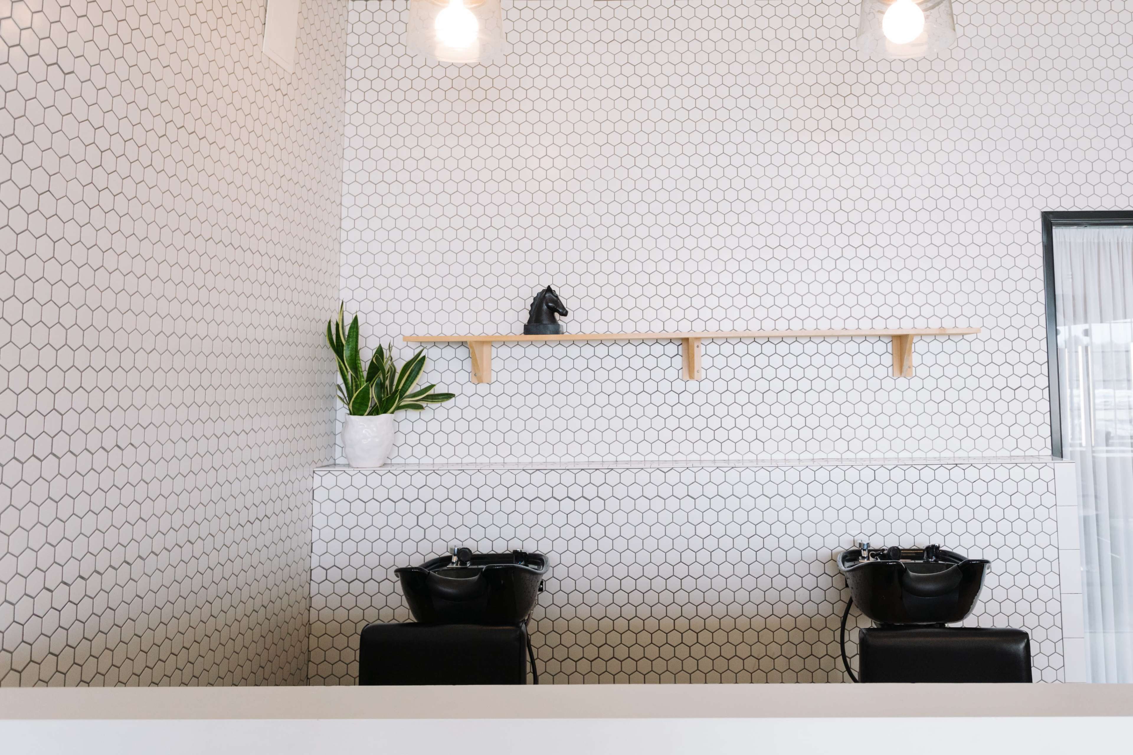 A minimalist hair salon with a hexagonal tiled wall, a shelf of wood, and two black shampoo bowls.