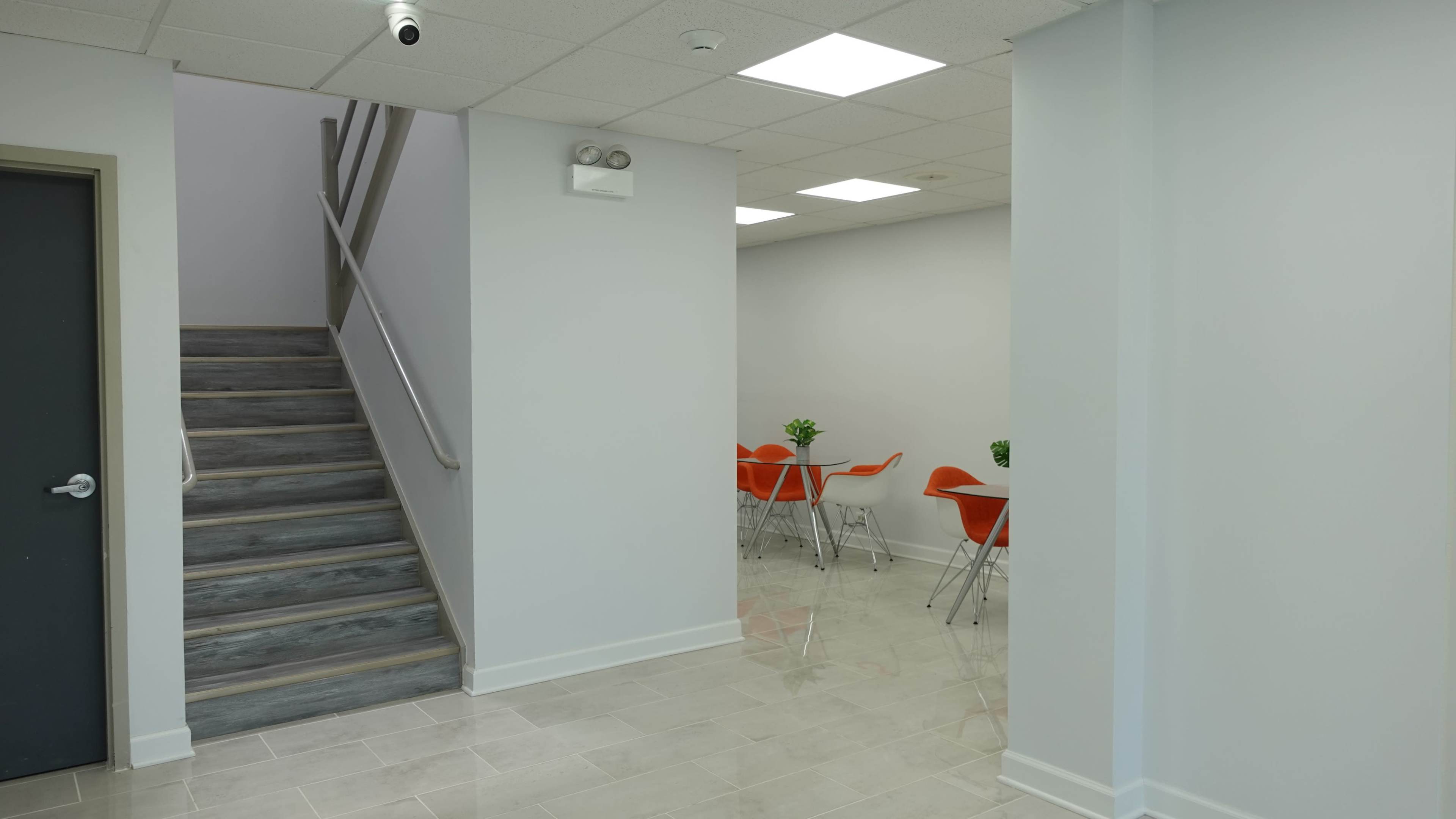 The image shows an interior space featuring a staircase on the left and a small dining area with orange chairs in the background.