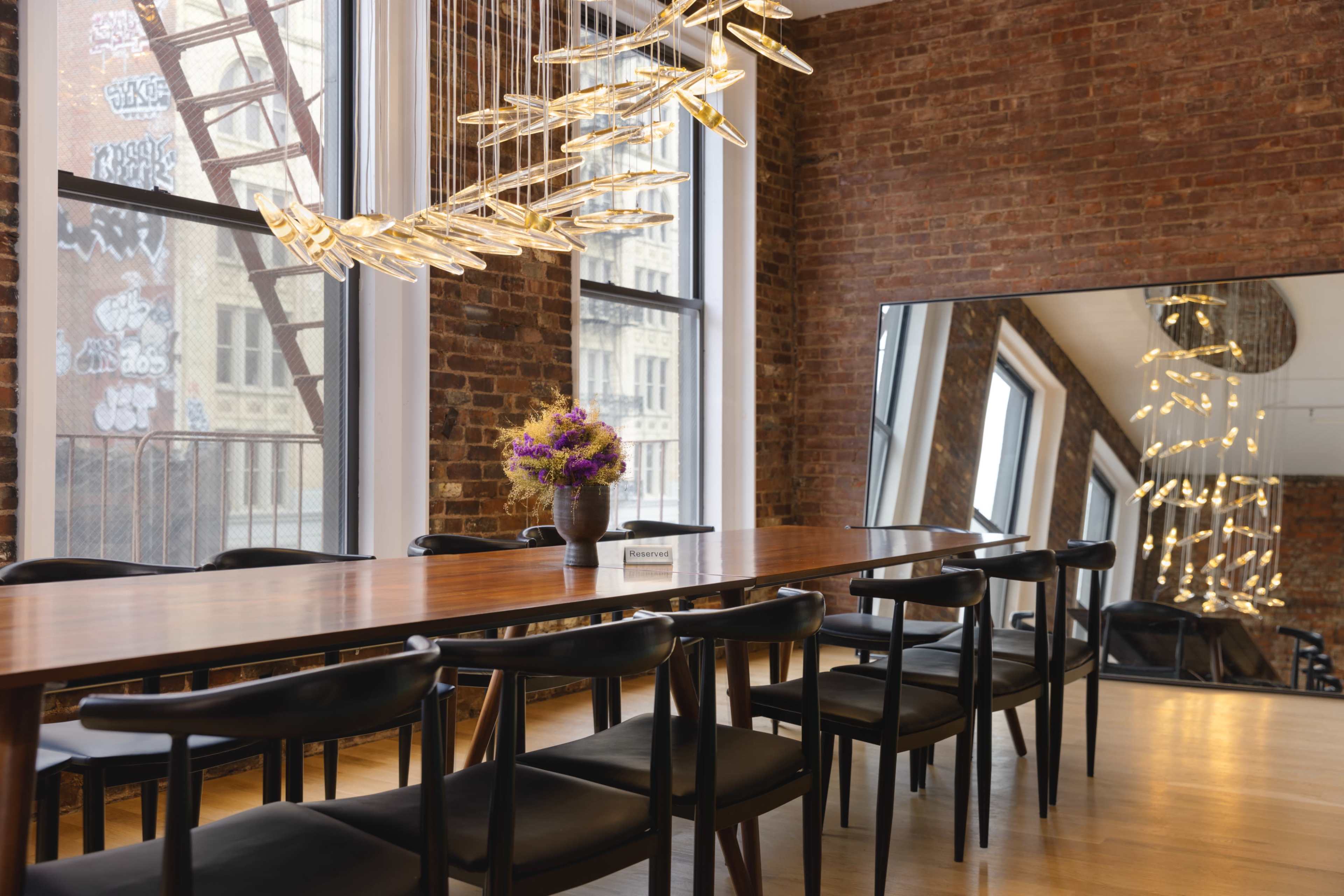 A long wooden dining table with black chairs is set in a room with exposed brick walls, large windows, and a modern light fixture above.