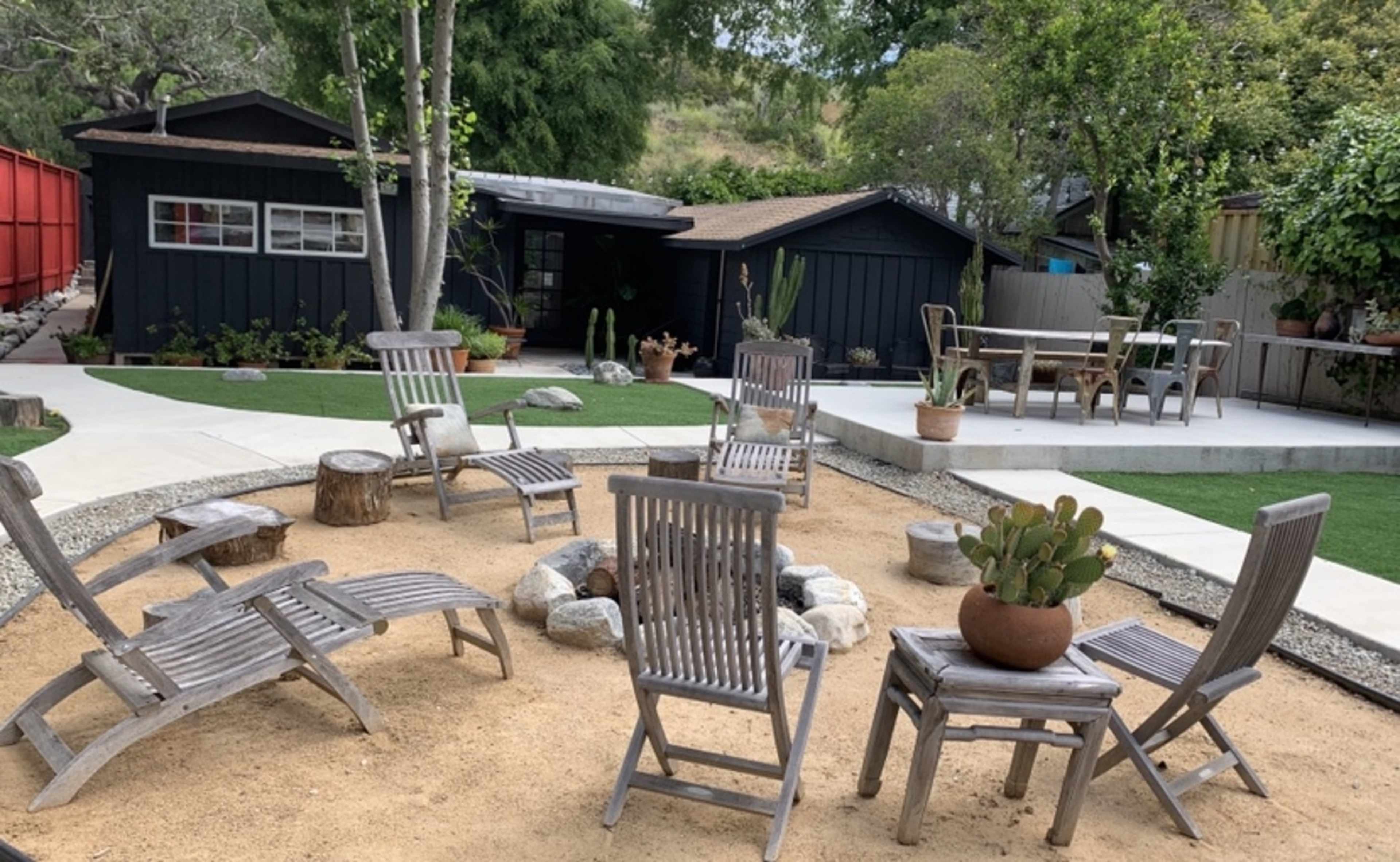 A landscaped outdoor area with wooden chairs arranged around a sandy space, surrounded by rocks, potted plants, and two structures in the background.