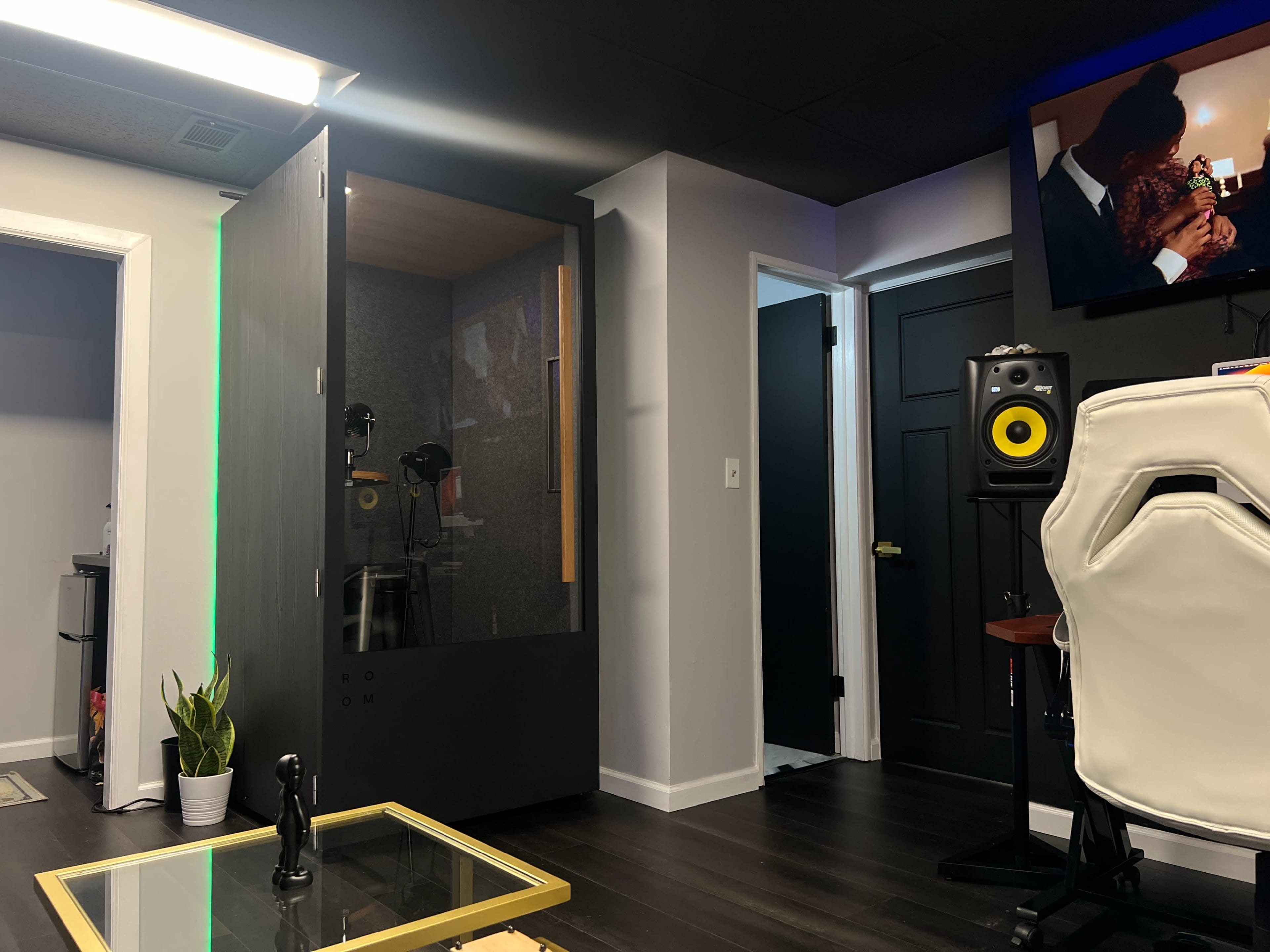 A modern recording studio with a soundproof booth, a computer desk, and a television on the wall.
