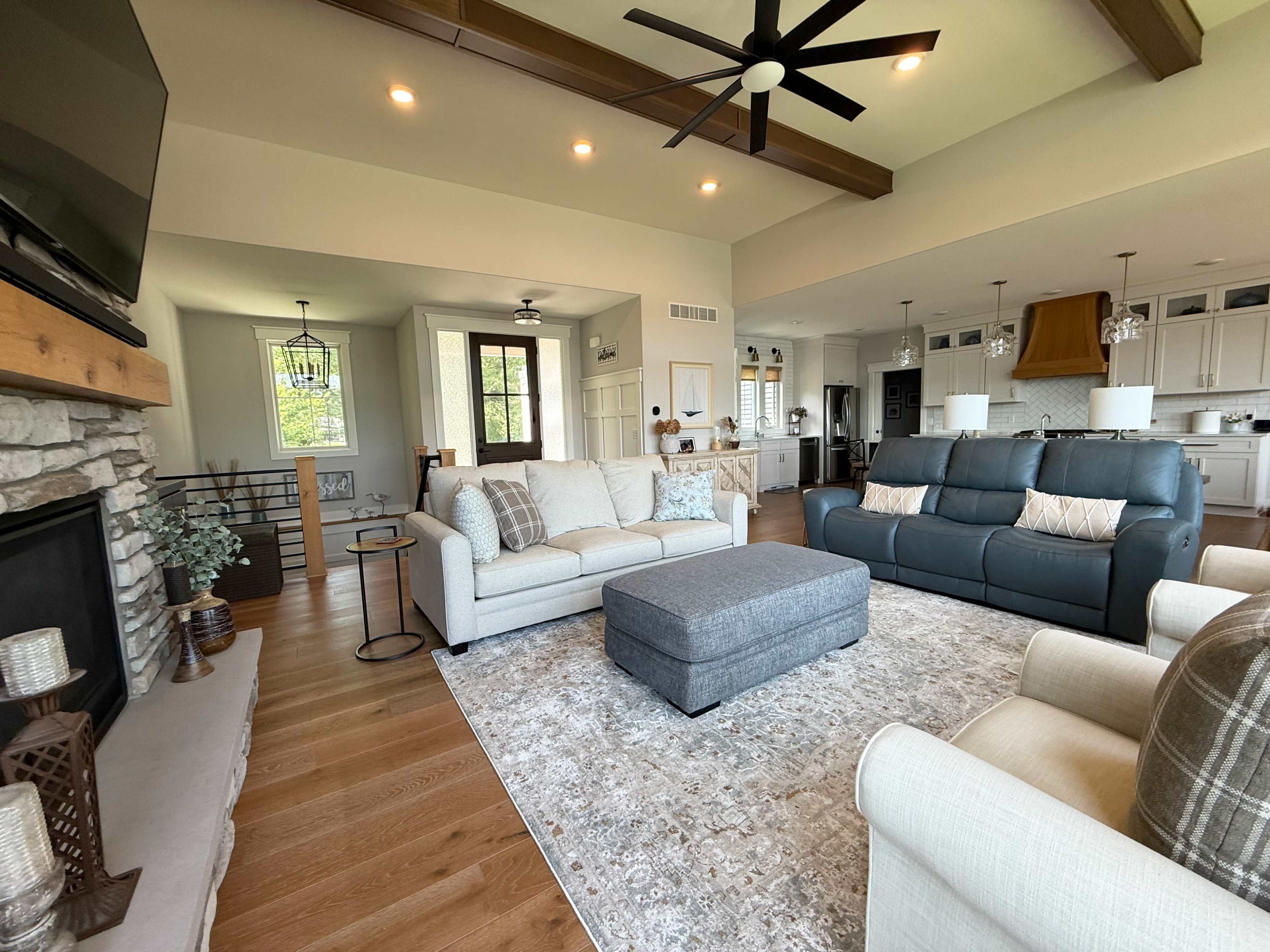 The living room features a light-colored sectional sofa, a dark gray ottoman, and a stone fireplace, with large windows providing natural light.