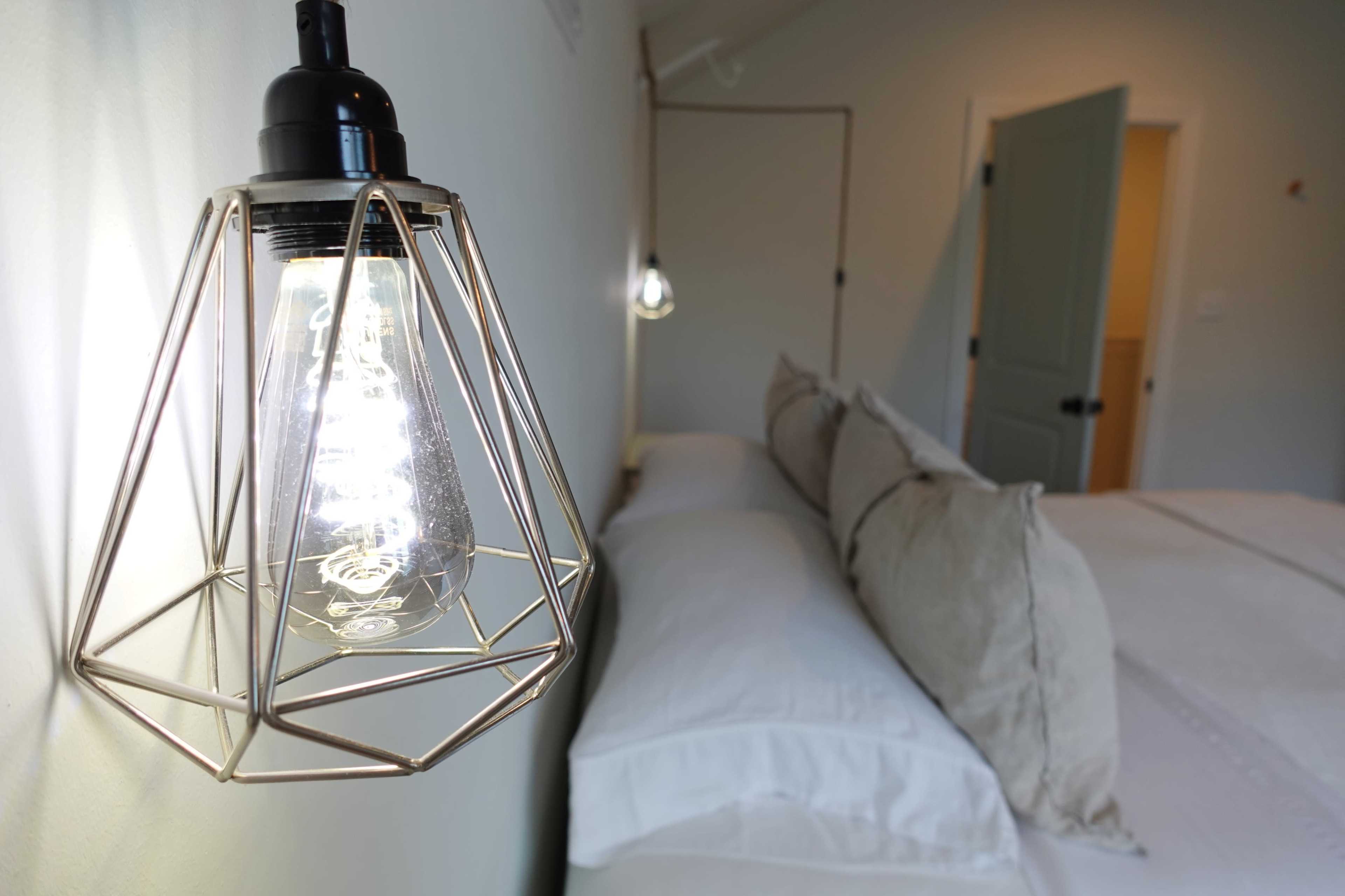 A pendant light with a metal cage hangs next to a neatly made bed featuring white linens and decorative pillows.