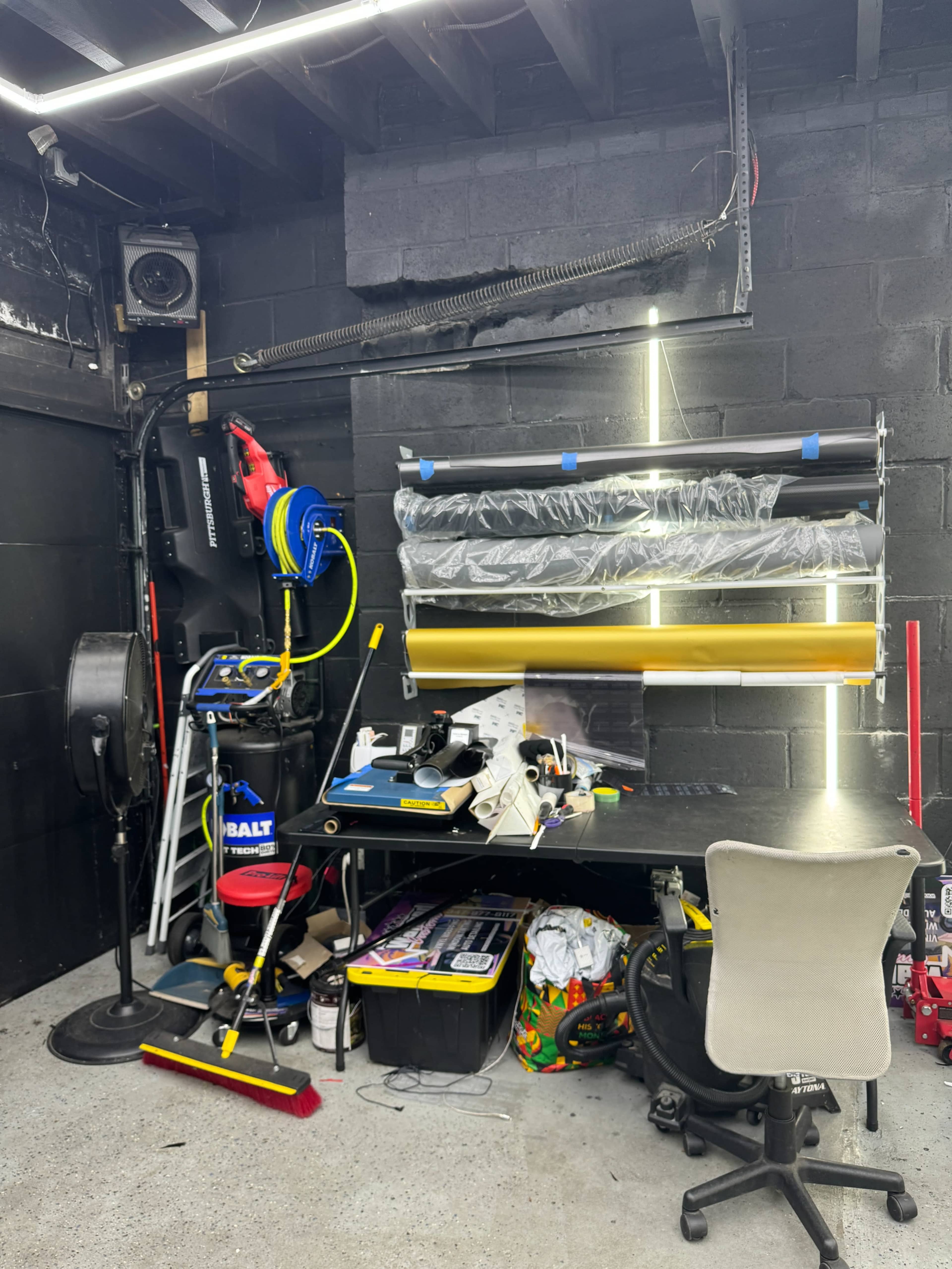 A workshop corner with a workbench, various tools, a roll holder for materials, and a chair amid a clutter of equipment and supplies.