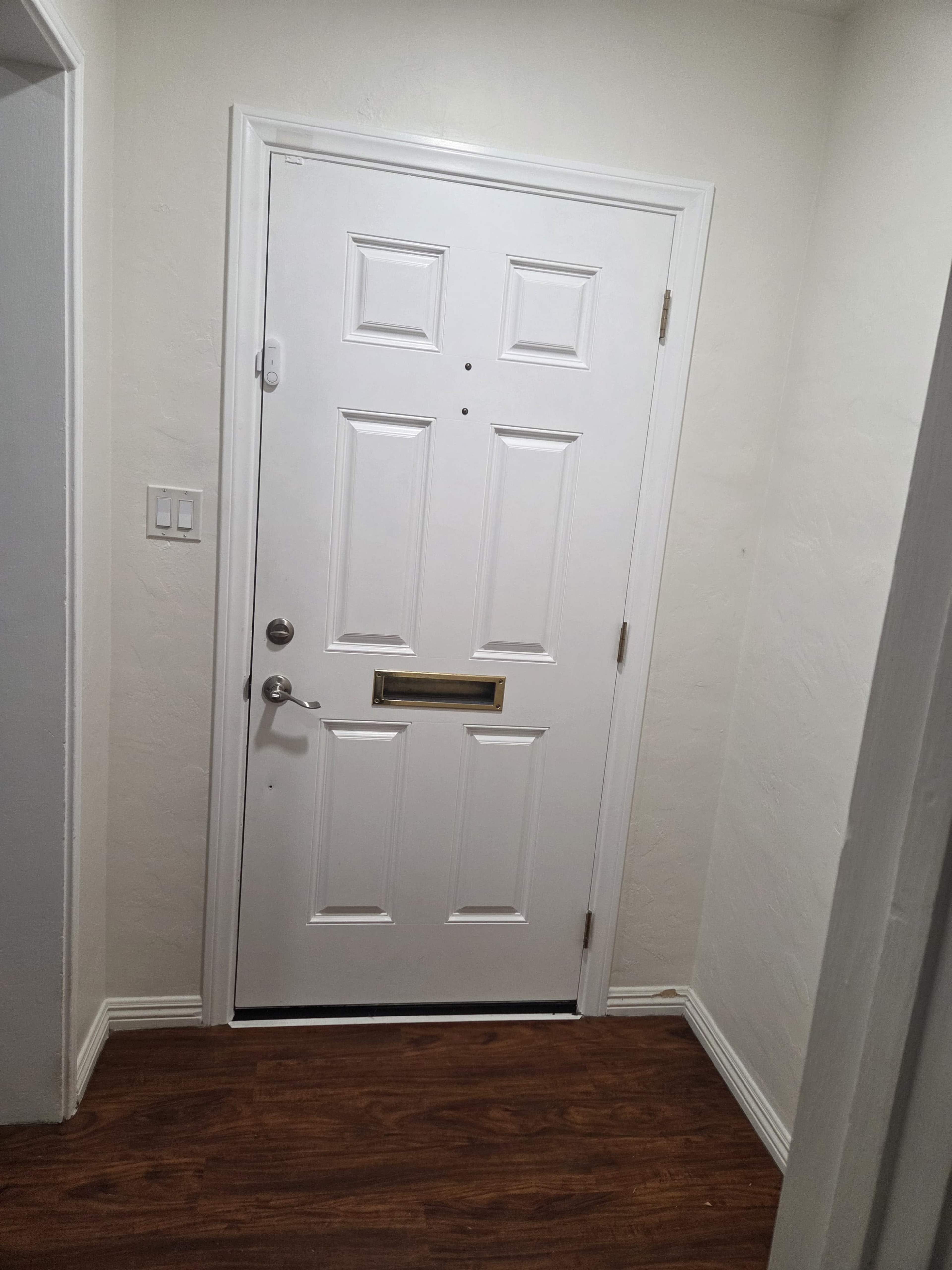 The image shows a white front door with a mail slot, situated in a hallway with light-colored walls and wooden flooring.
