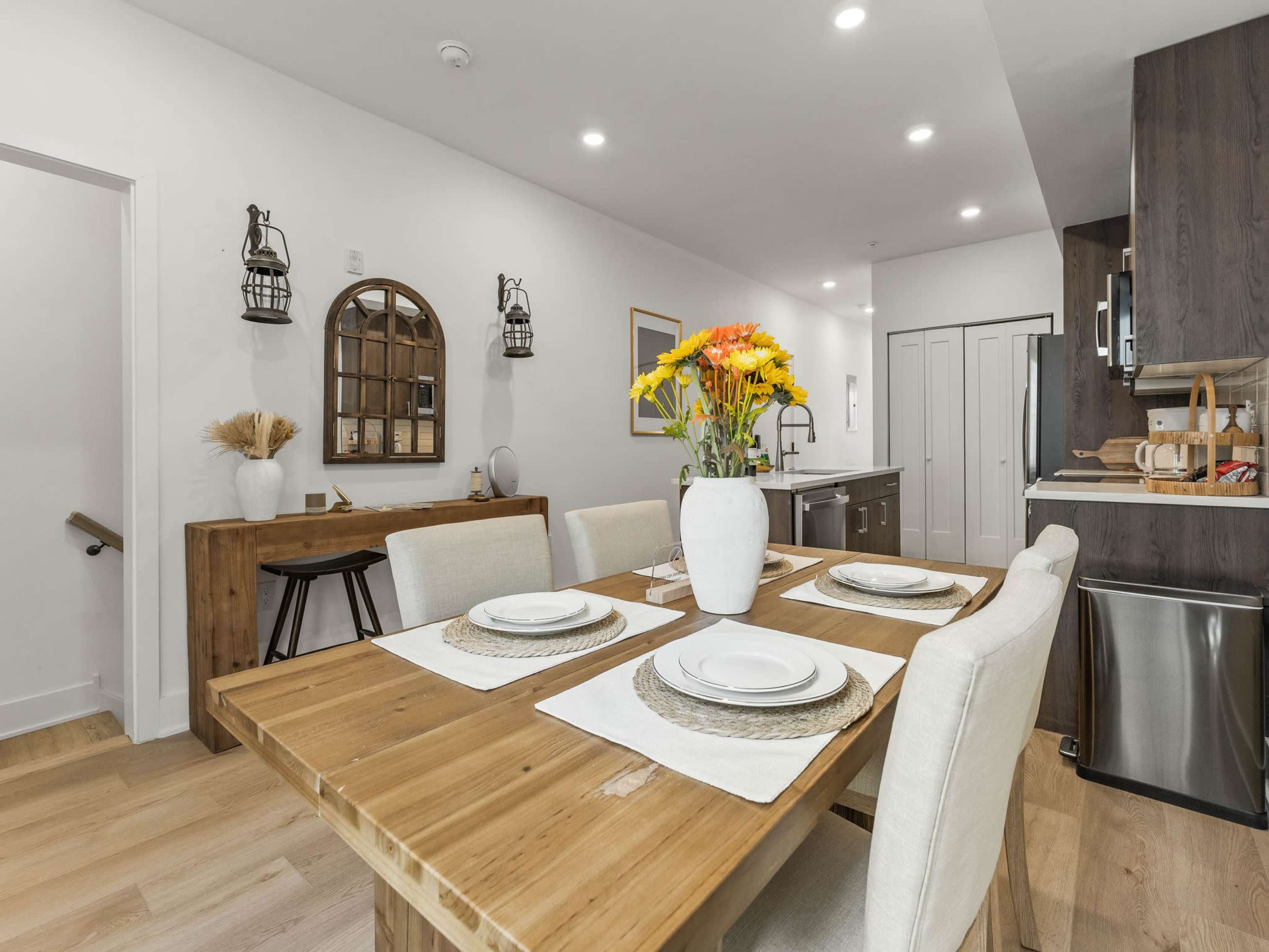 The image shows a modern dining area with a wooden table set for a meal, featuring a vase of flowers, and a kitchen visible in the background.