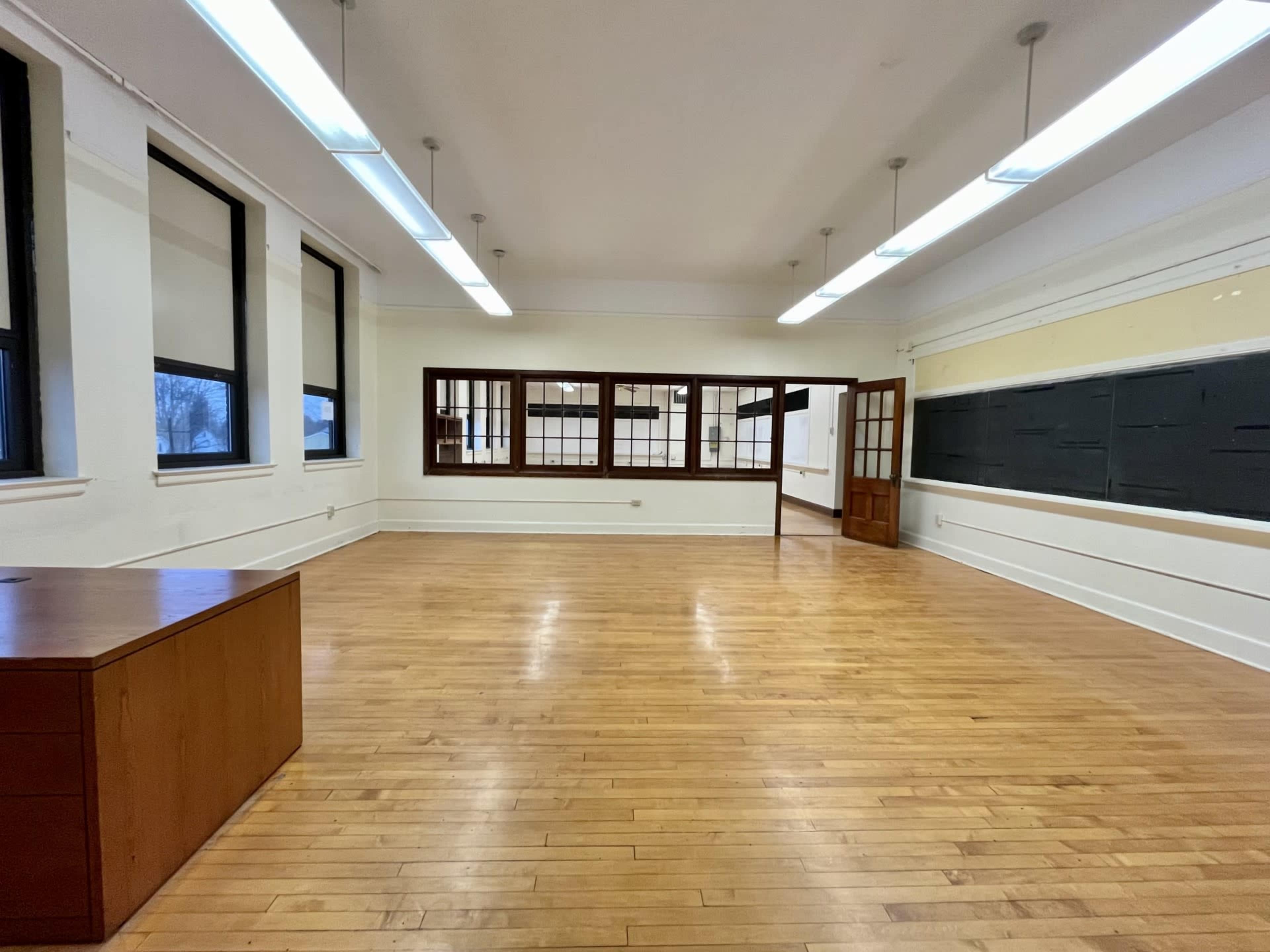 The image shows a spacious, empty classroom with wooden floors, large windows, and a door leading to another room.