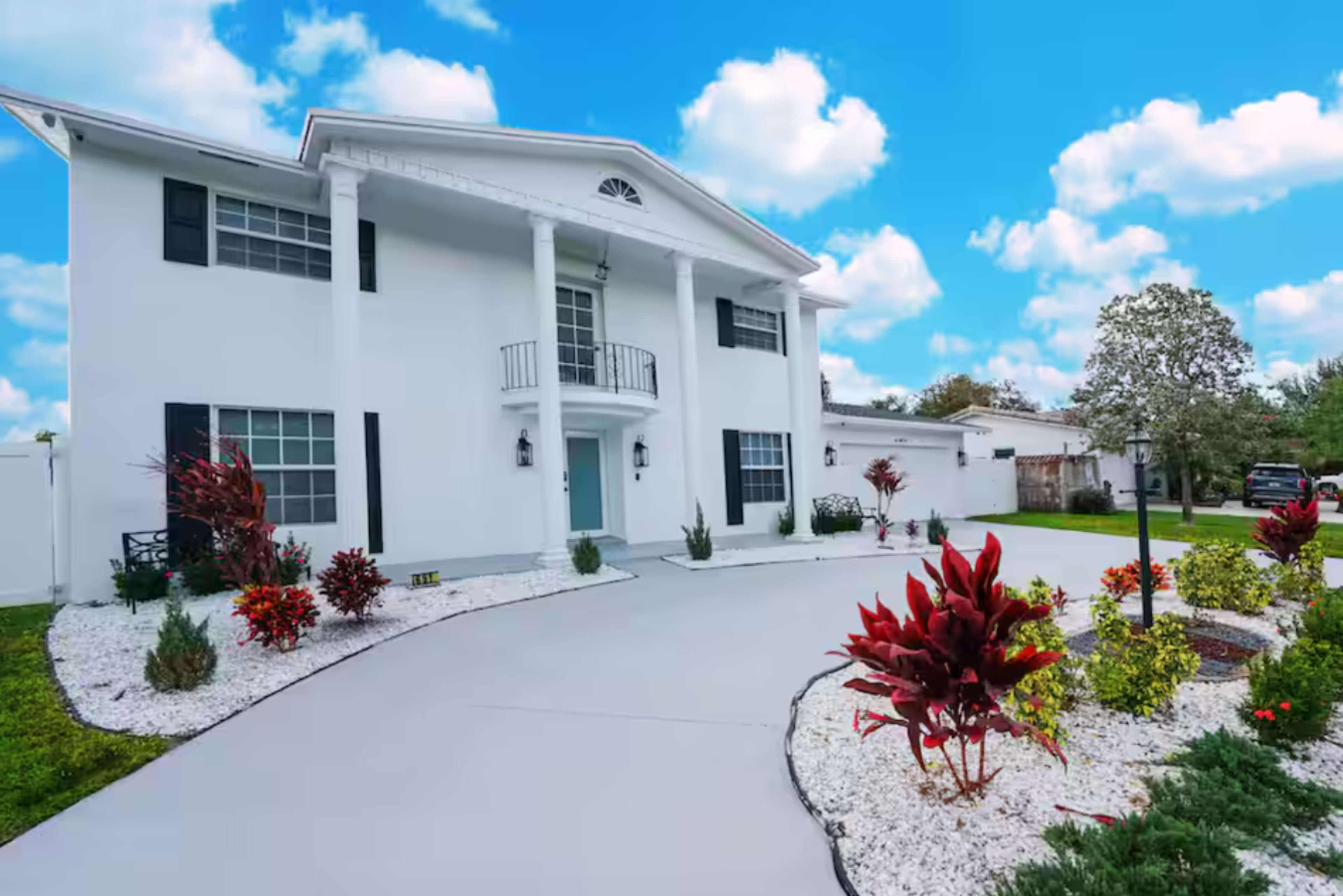 A two-story white house with black shutters features a landscaped front yard with colorful plants and a curved driveway.