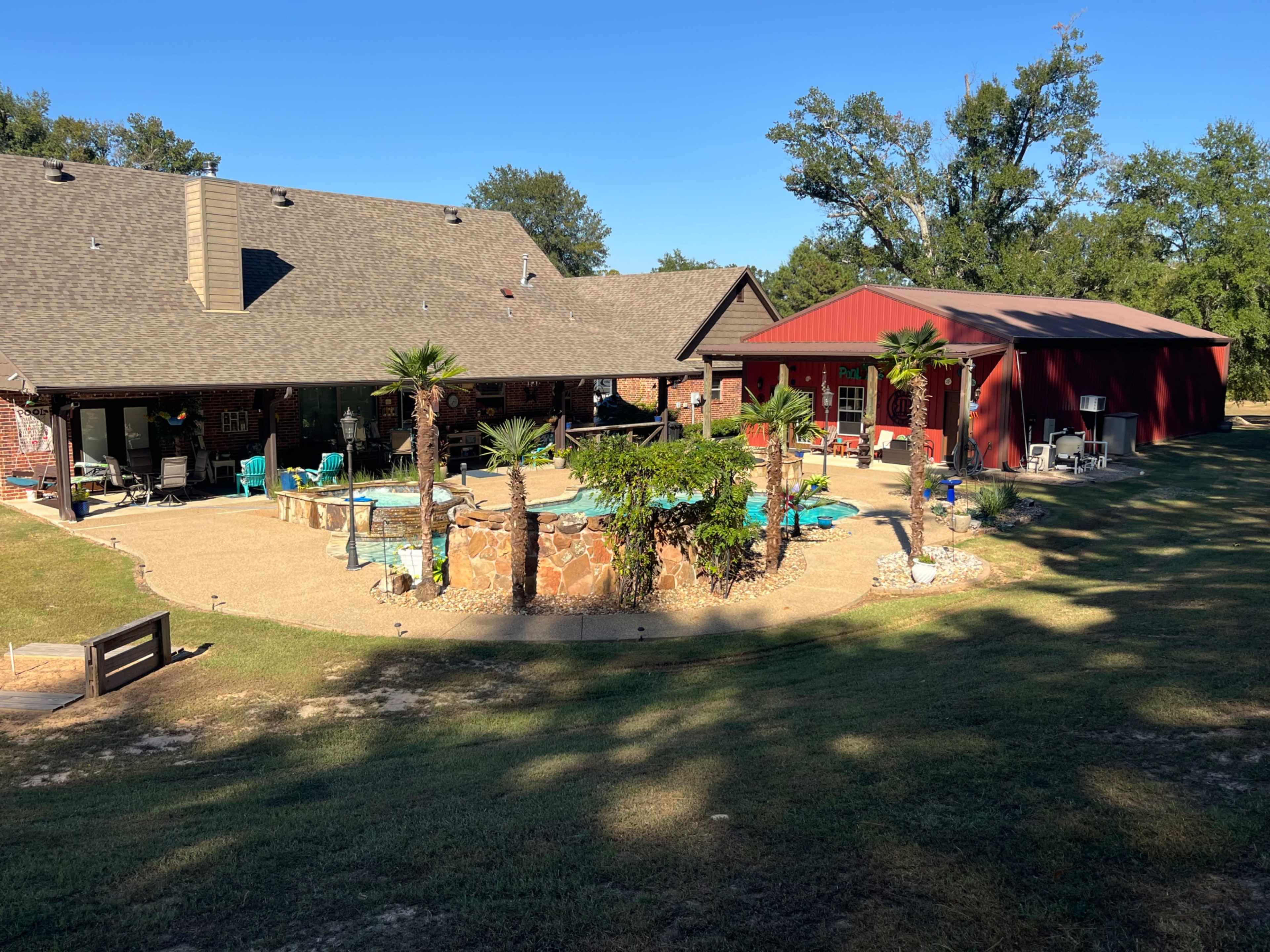 The image shows a backyard scene featuring a landscaped area with a pool, surrounded by palm trees, and a large red shed adjacent to a brick house.