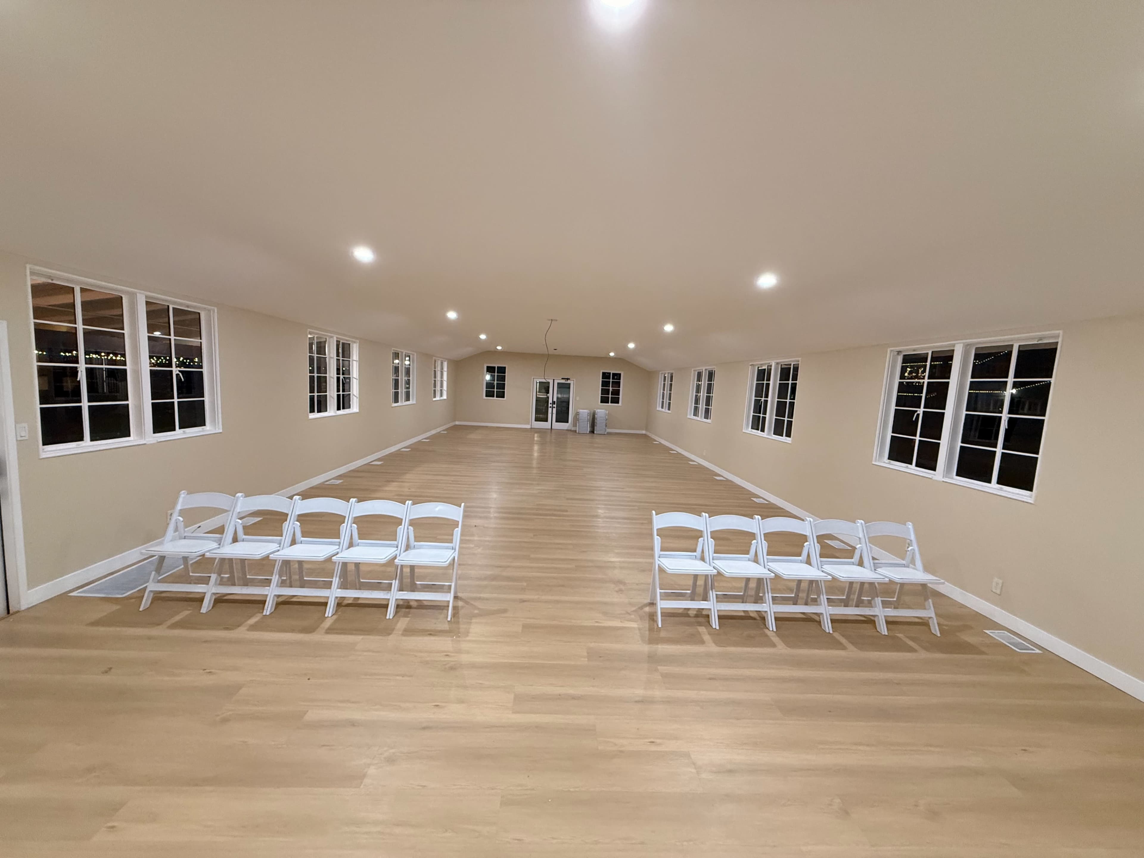 The image shows a large, empty room with wooden floors and several rows of white folding chairs set up facing an open space, illuminated by overhead lighting and lined with windows.