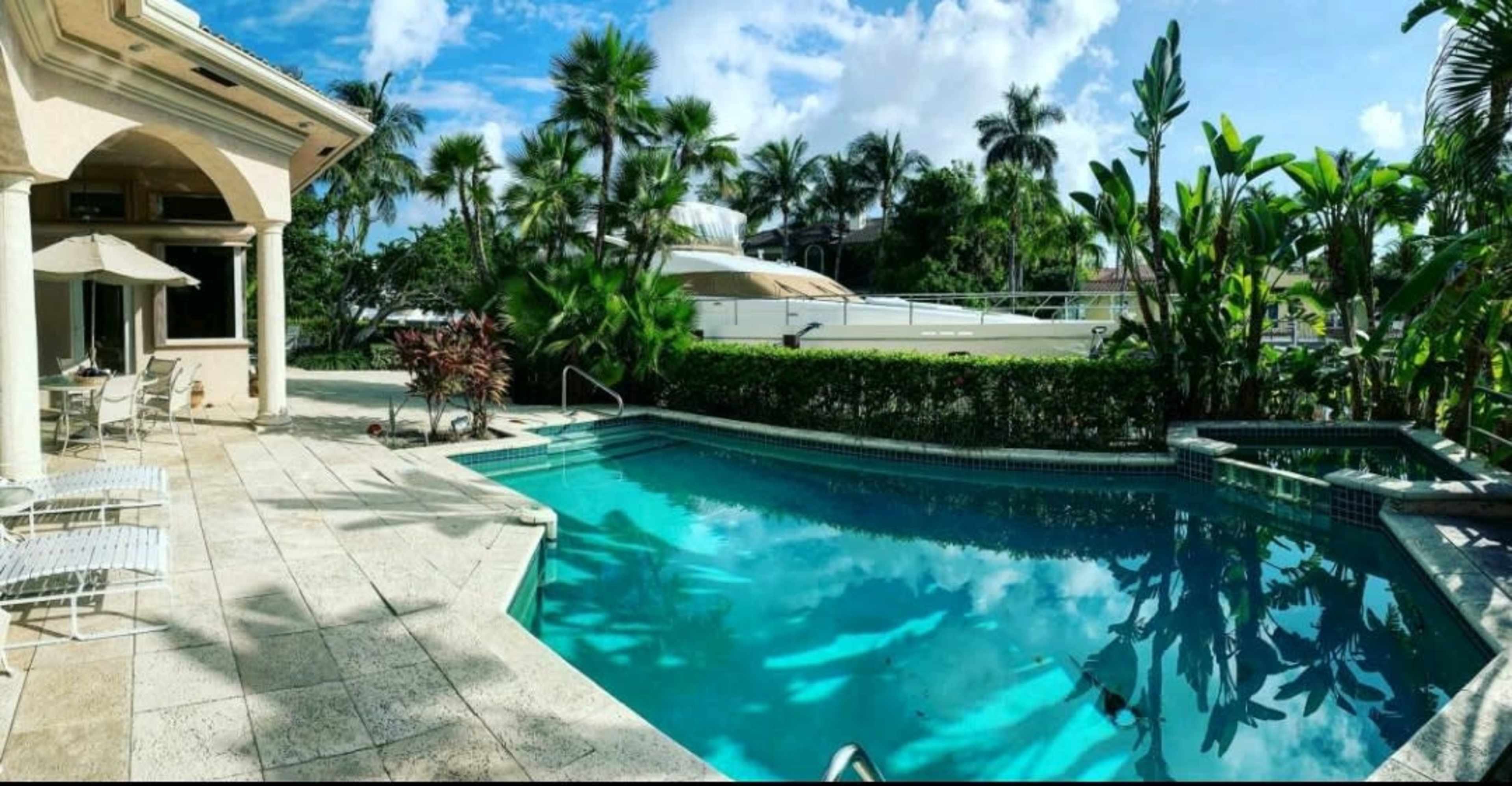A pool area surrounded by palm trees and tropical plants, with a patio and seating arrangement beside the water.