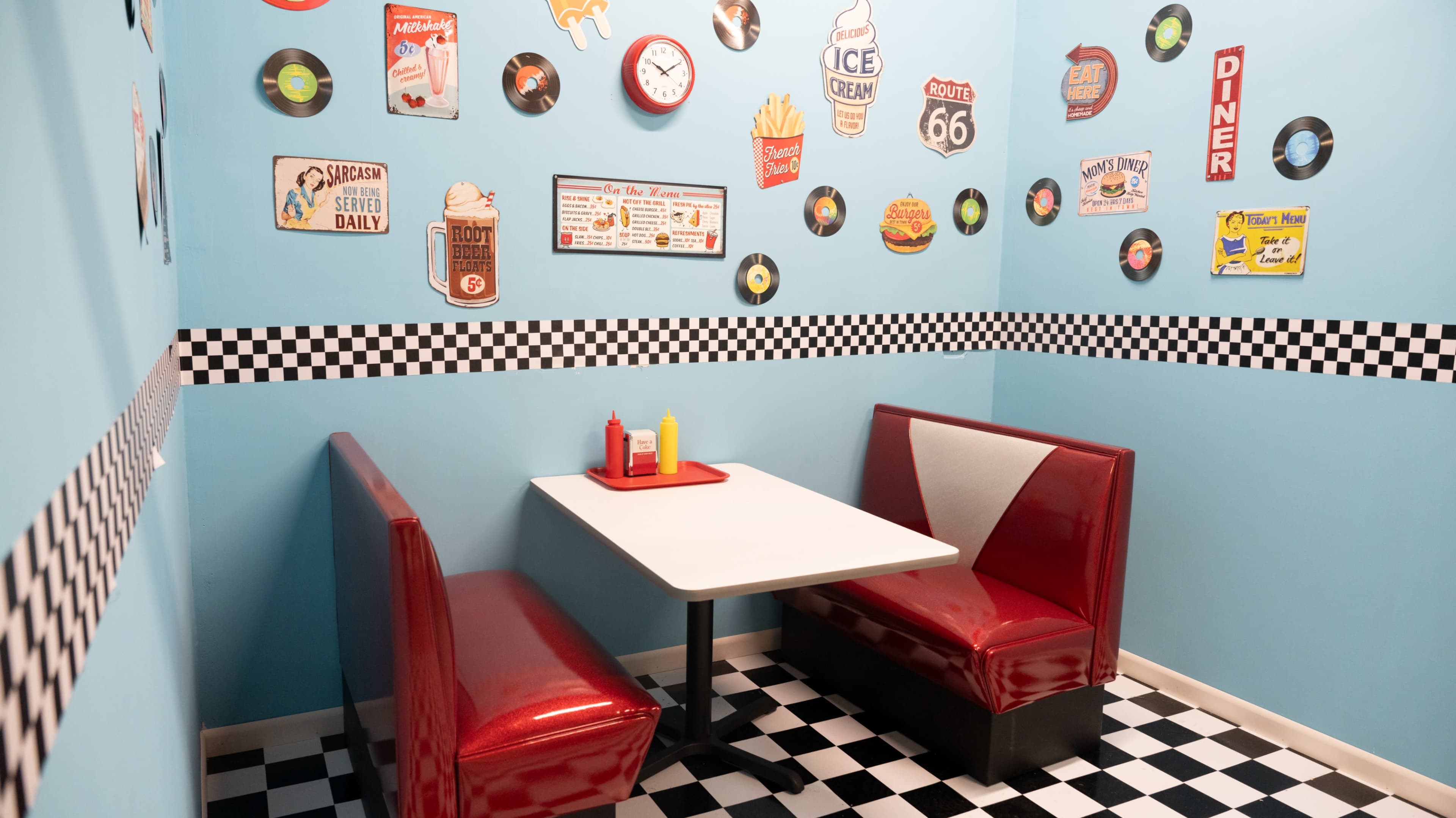 The image shows a retro diner booth with red seating, a white table, and walls decorated with vintage signs and records in a checkered pattern.