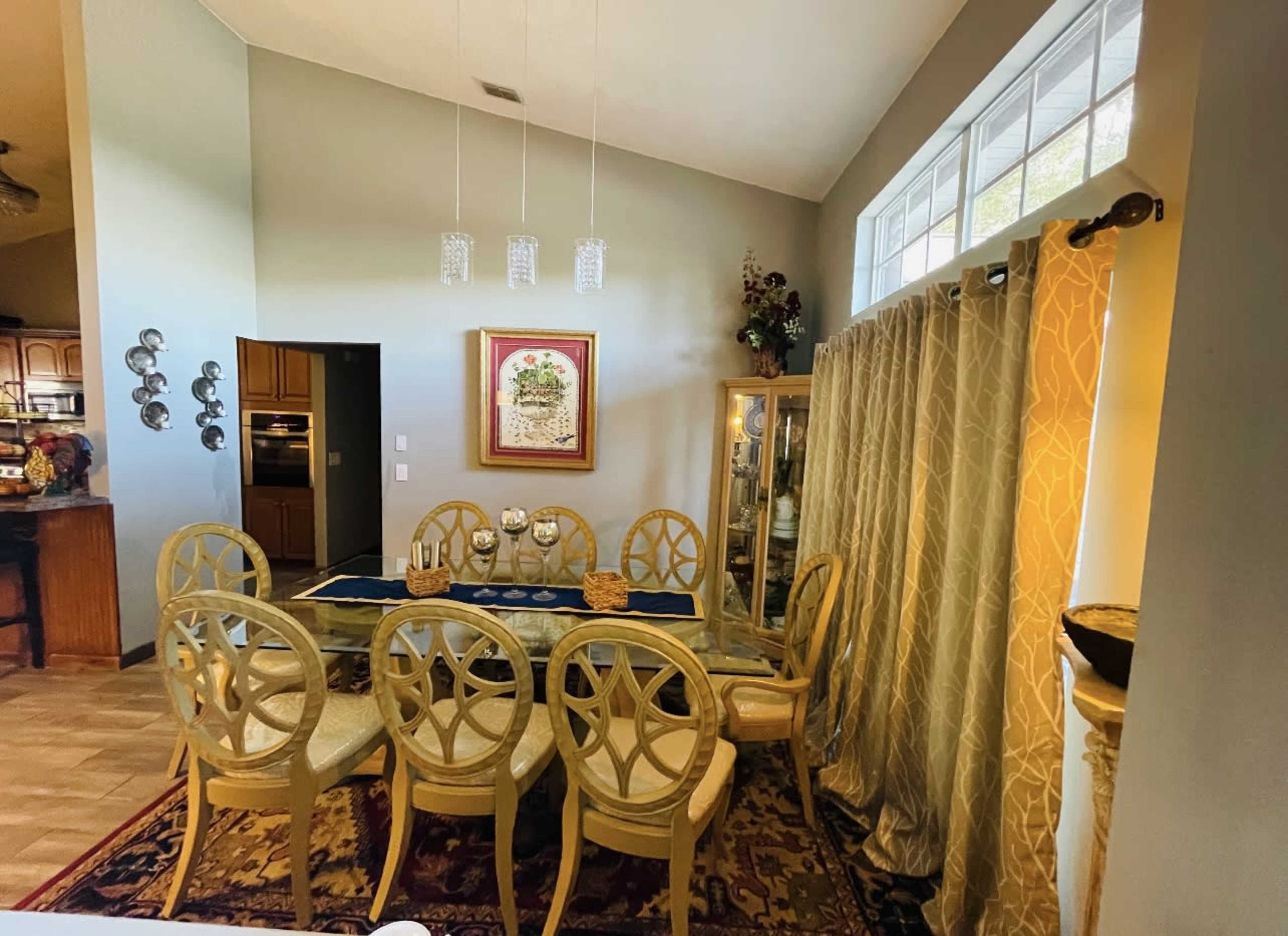 A dining area features a large table surrounded by eight chairs, with a nearby window and a decorative cabinet against the wall.