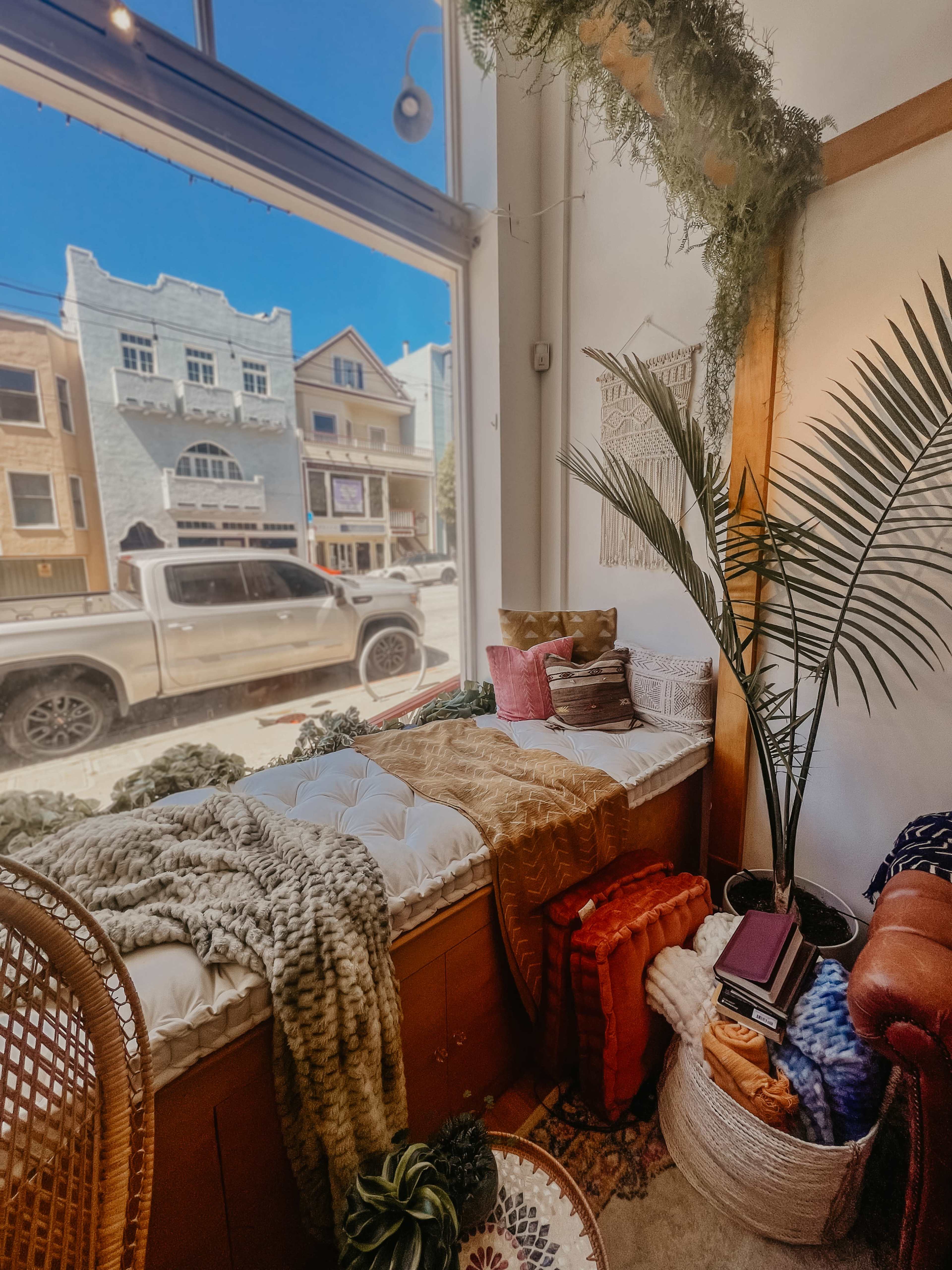 A cozy window seat adorned with blankets and decorative pillows overlooks a street with colorful buildings and a parked truck.
