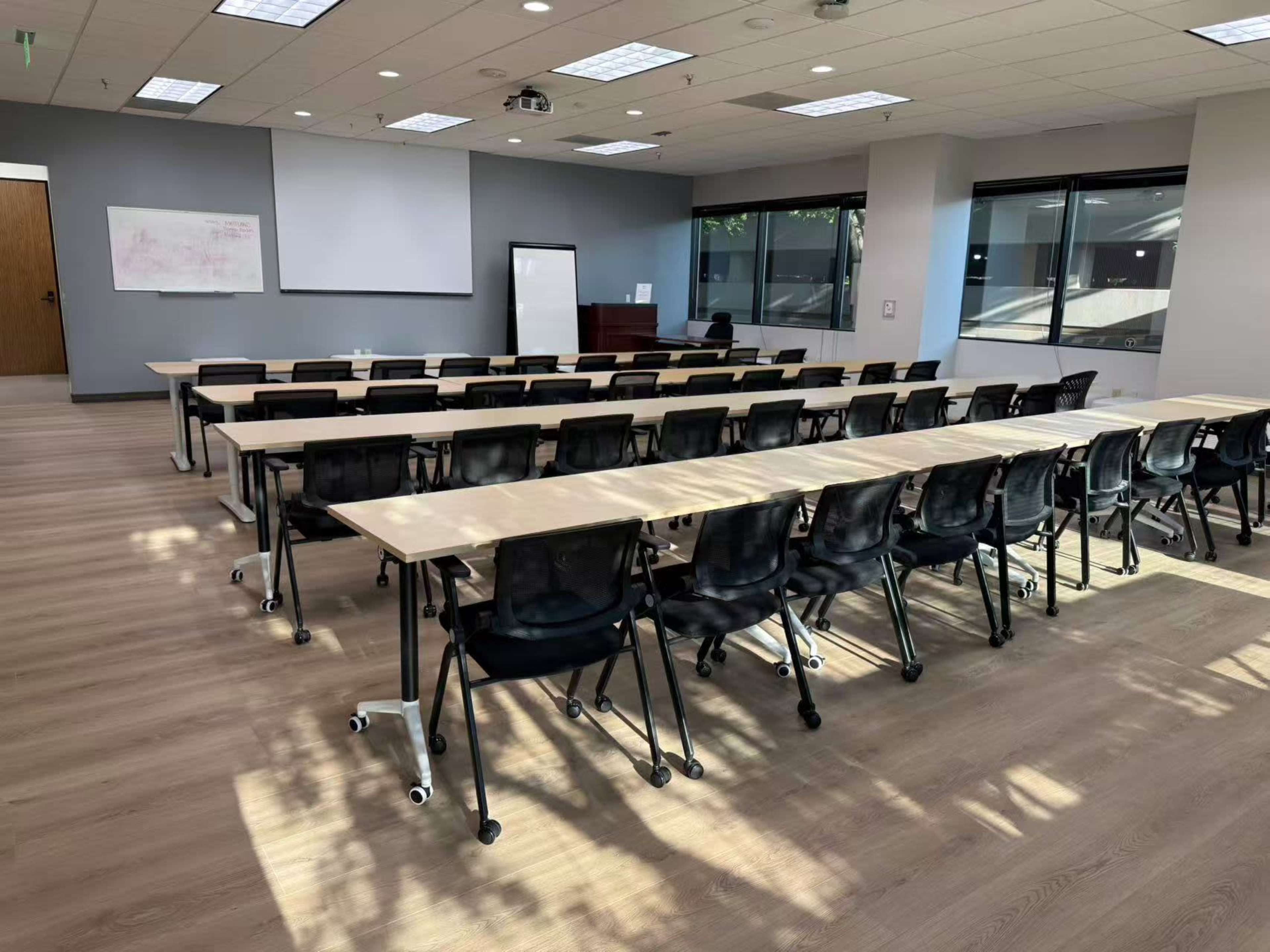 The image shows a classroom setup with multiple rows of tables and chairs, a whiteboard, and a projector in a well-lit room.