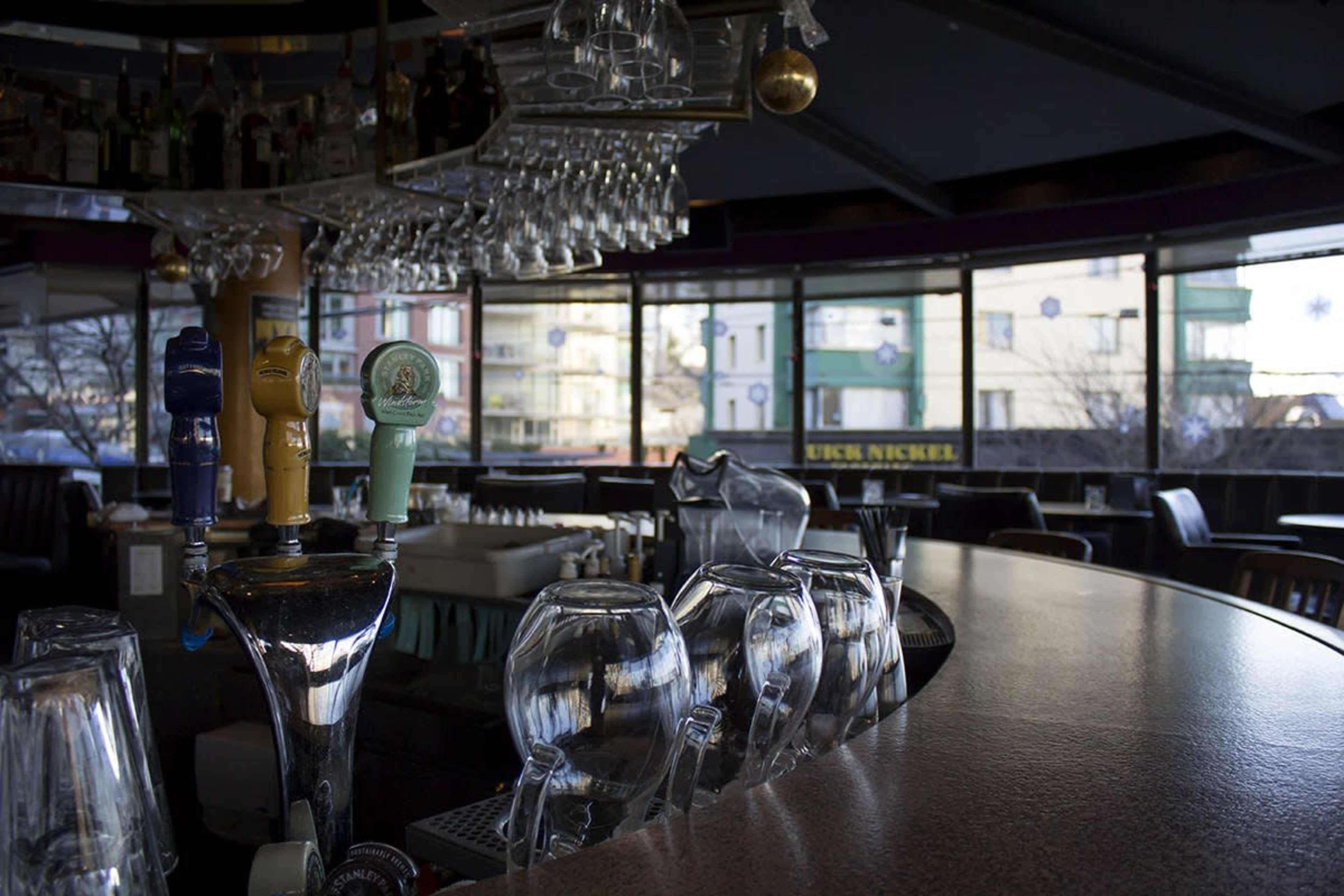 The scene shows a bar area with drink taps and a row of empty glasses, set against large windows featuring a city view.