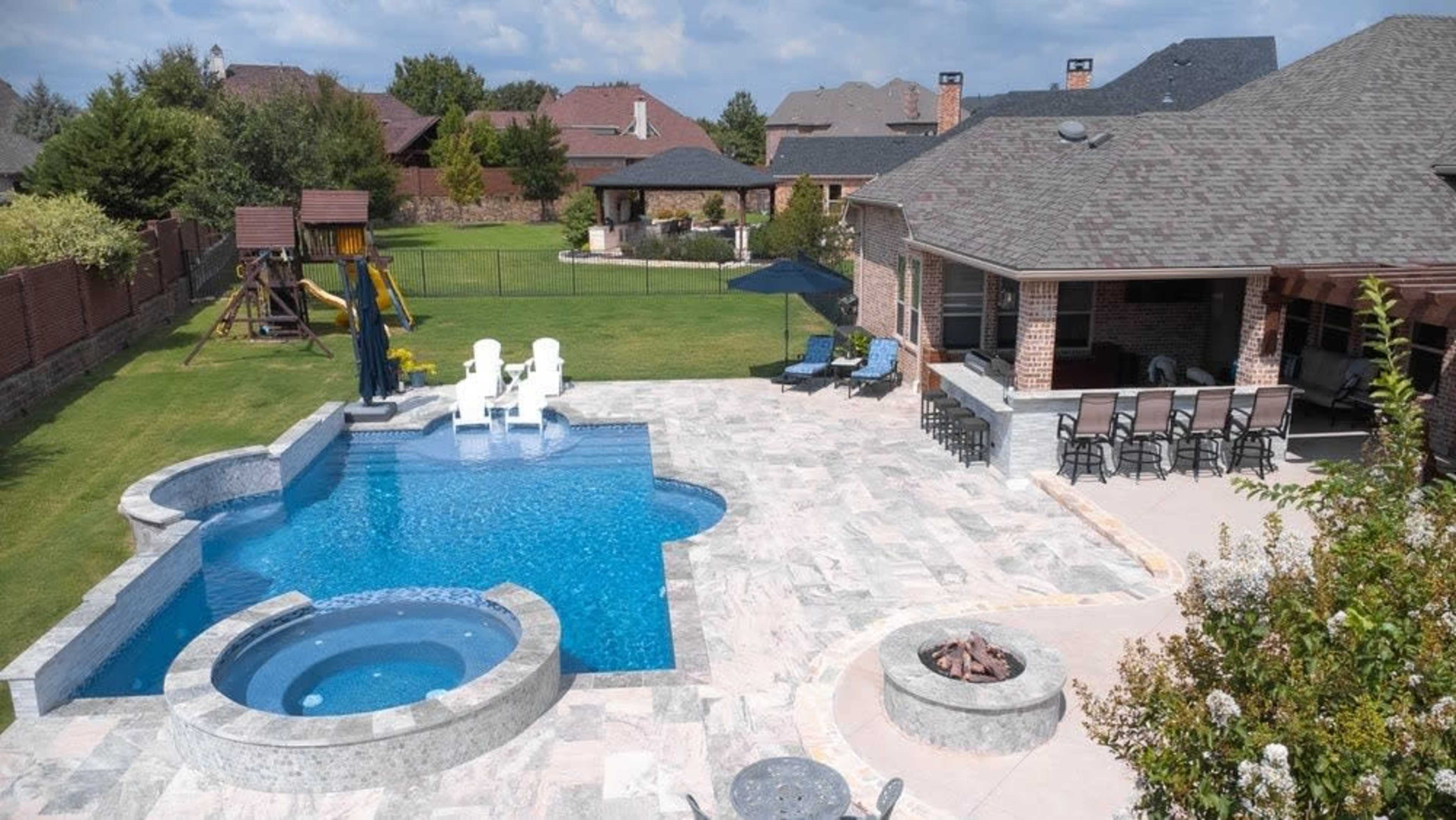 The image shows a landscaped backyard with a swimming pool, a spa, and an outdoor seating area beside a large house.