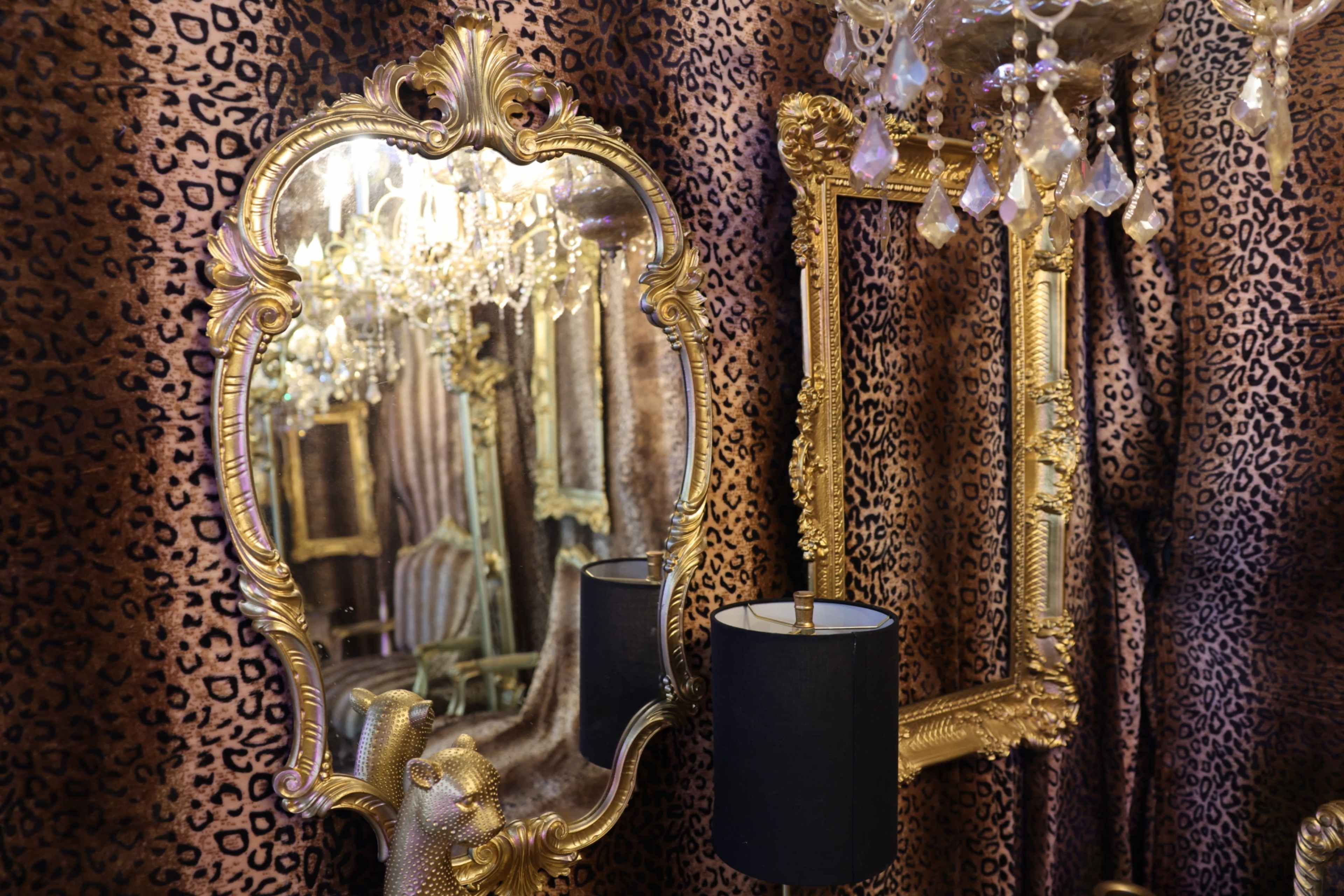 The image features ornate gold-framed mirrors alongside a black lamp, all set against a leopard-print backdrop with sparkling chandeliers.