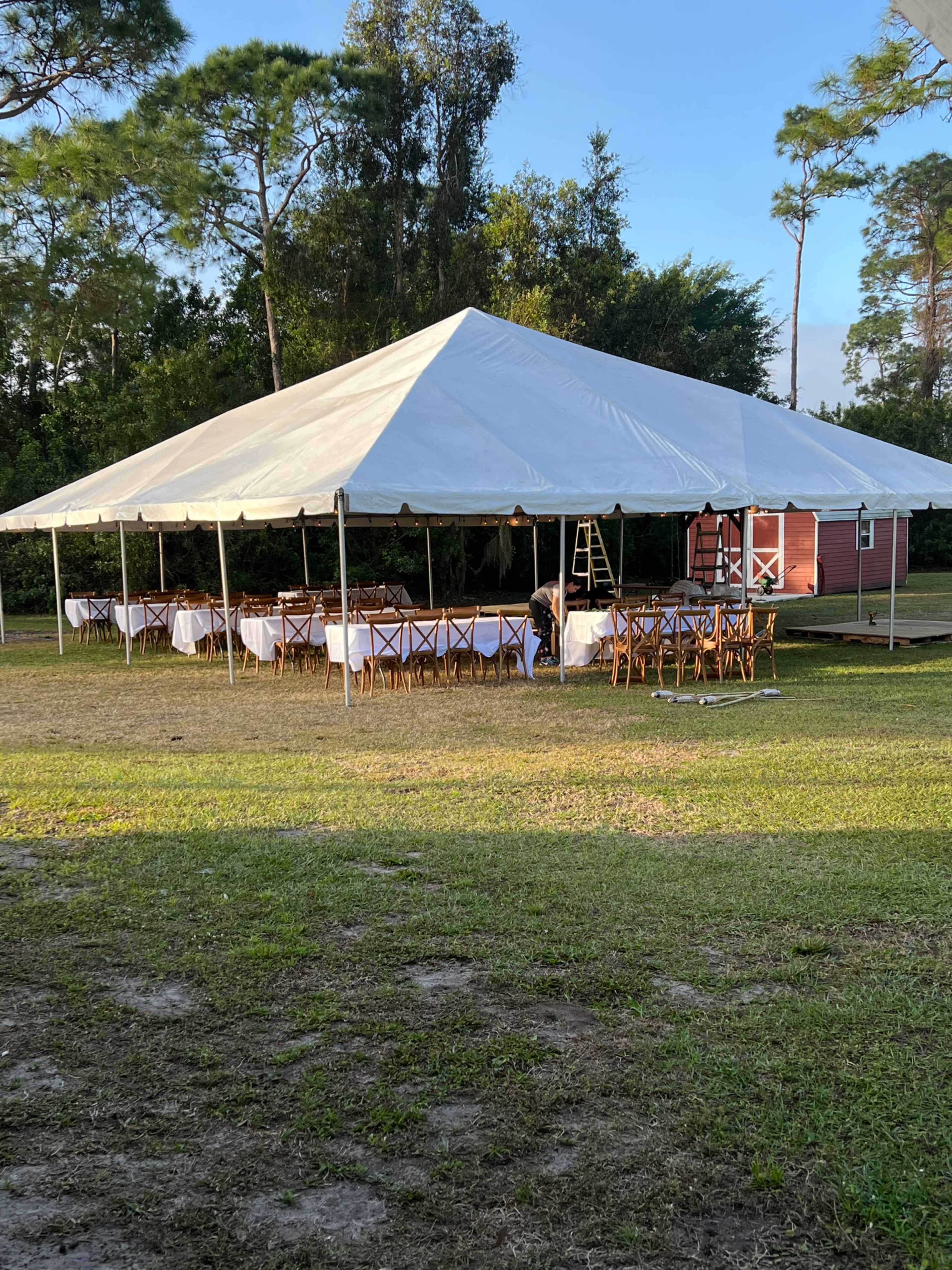 A large white tent is set up in a grassy area, surrounded by trees, with tables arranged underneath and a ladder leaning against one side.