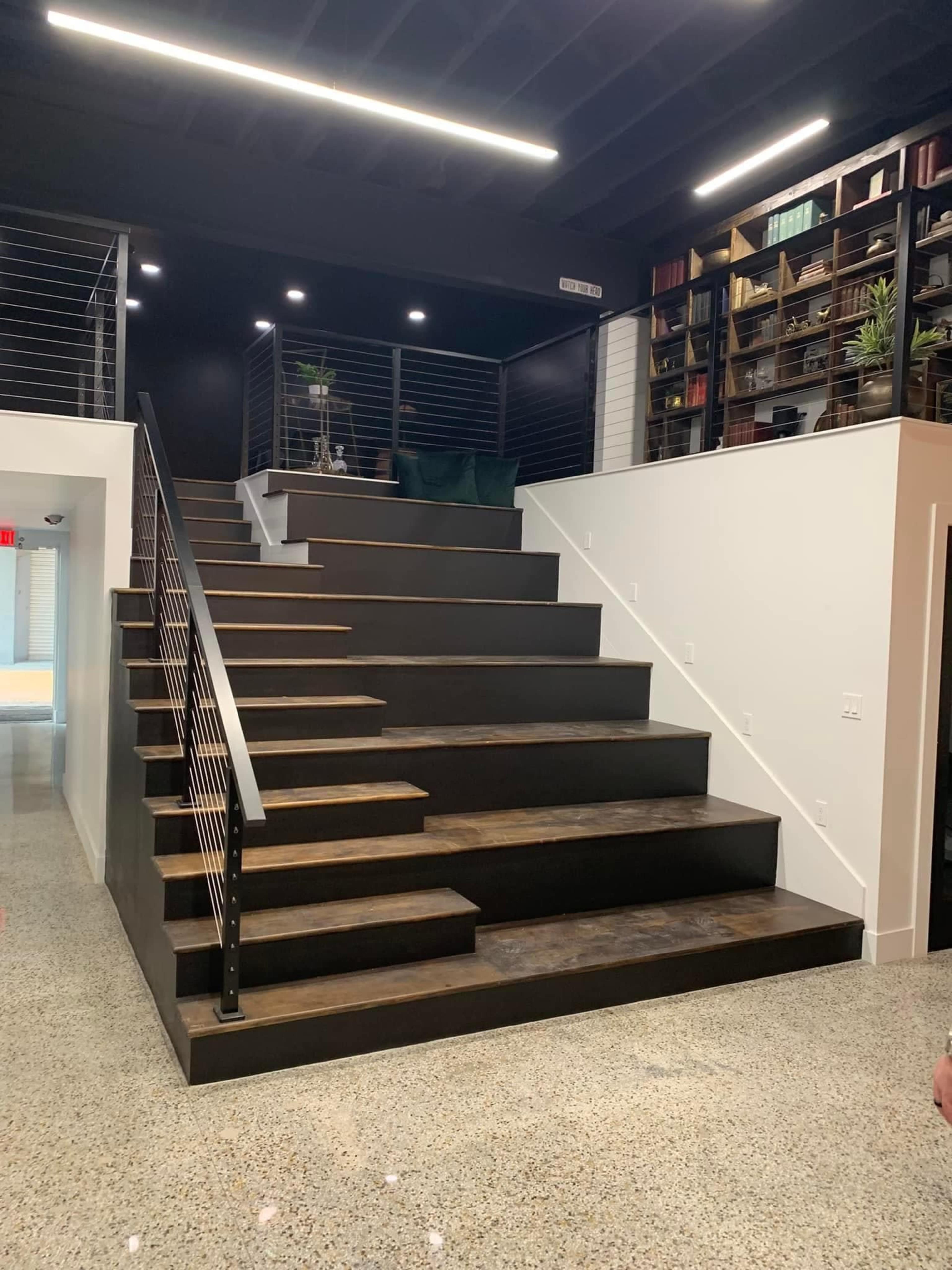 A modern staircase with dark wooden steps leading to an upper level in a well-lit contemporary space, featuring a railing on one side and a bookshelf on the wall.