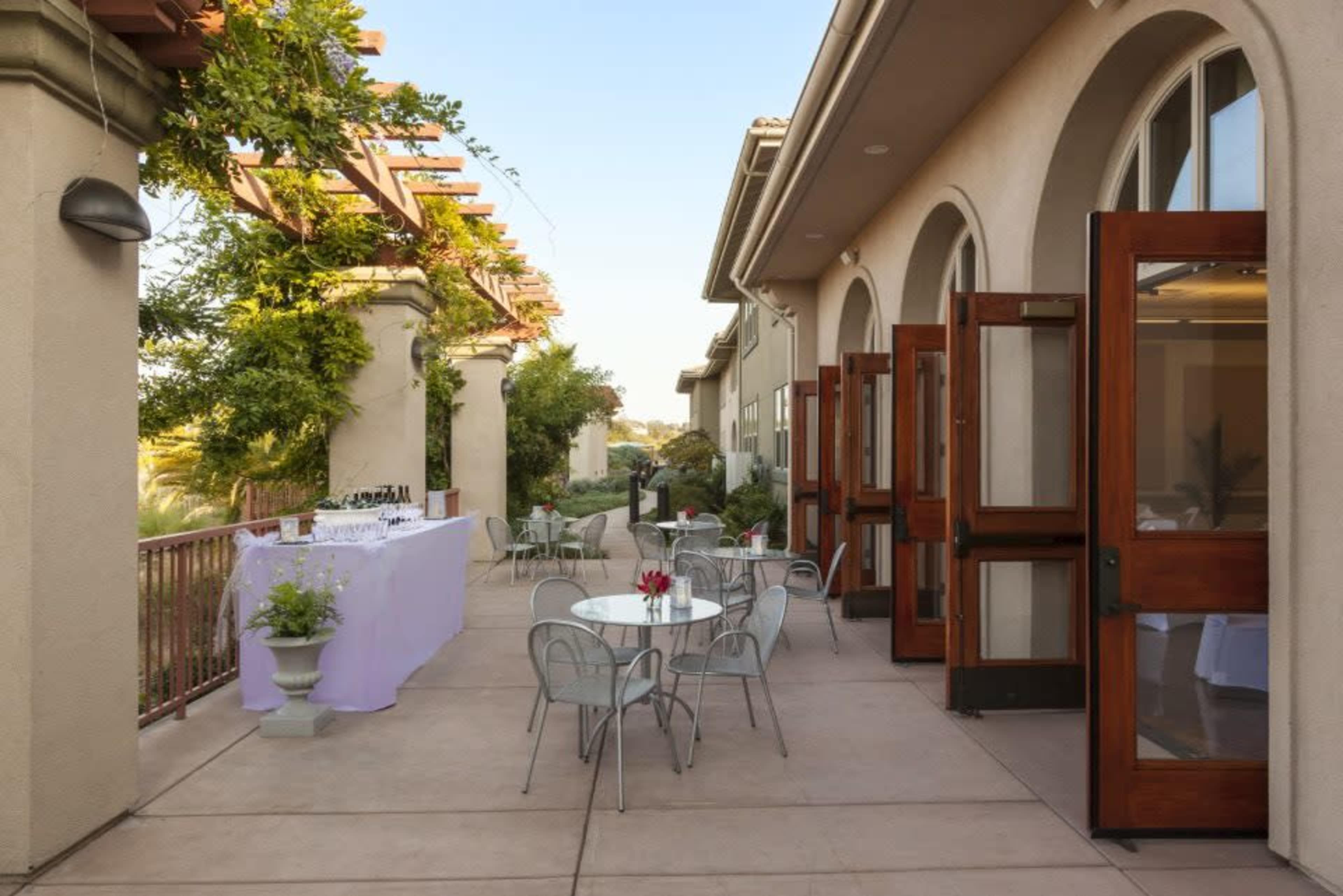 A patio area with tables and chairs, flanked by large arches and doors leading into a building, with a table set up for an event in the background.