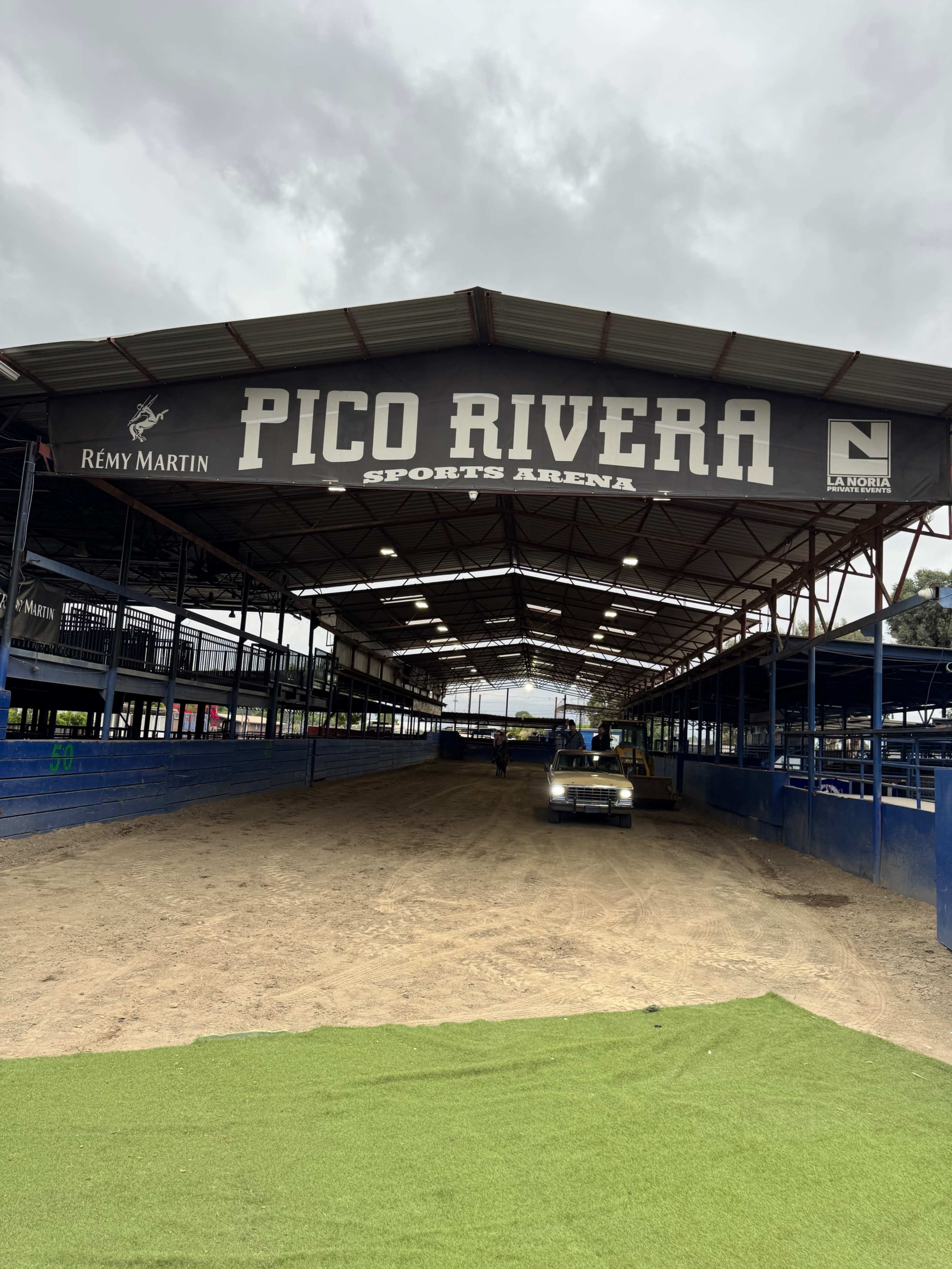 The image shows the interior of the Pico Rivera Sports Arena, featuring a dirt floor and covered seating areas under a large roof.