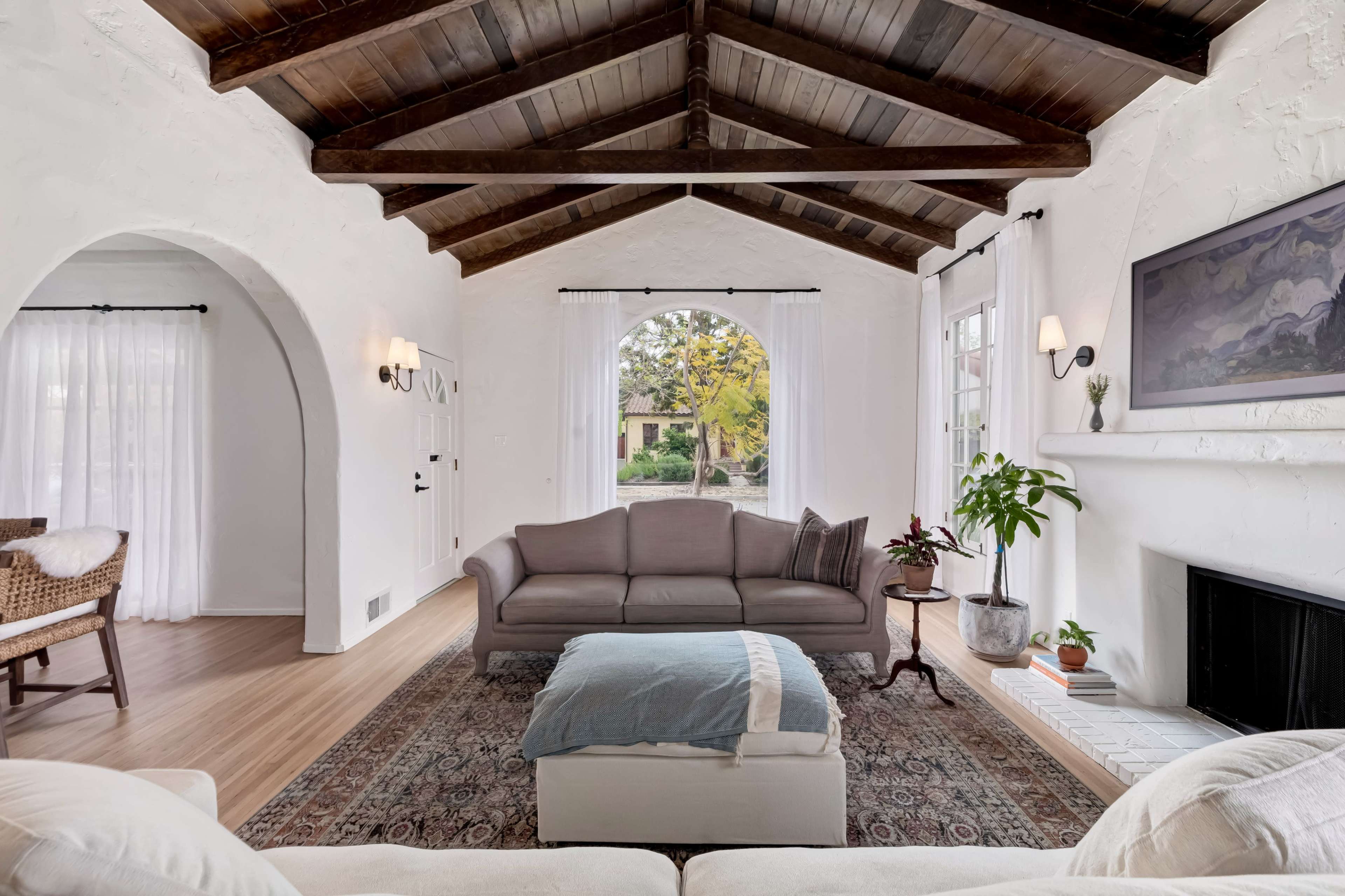 The image showcases a bright living room with vaulted wooden ceilings, a gray sofa facing a large window, and greenery accentuating the space.