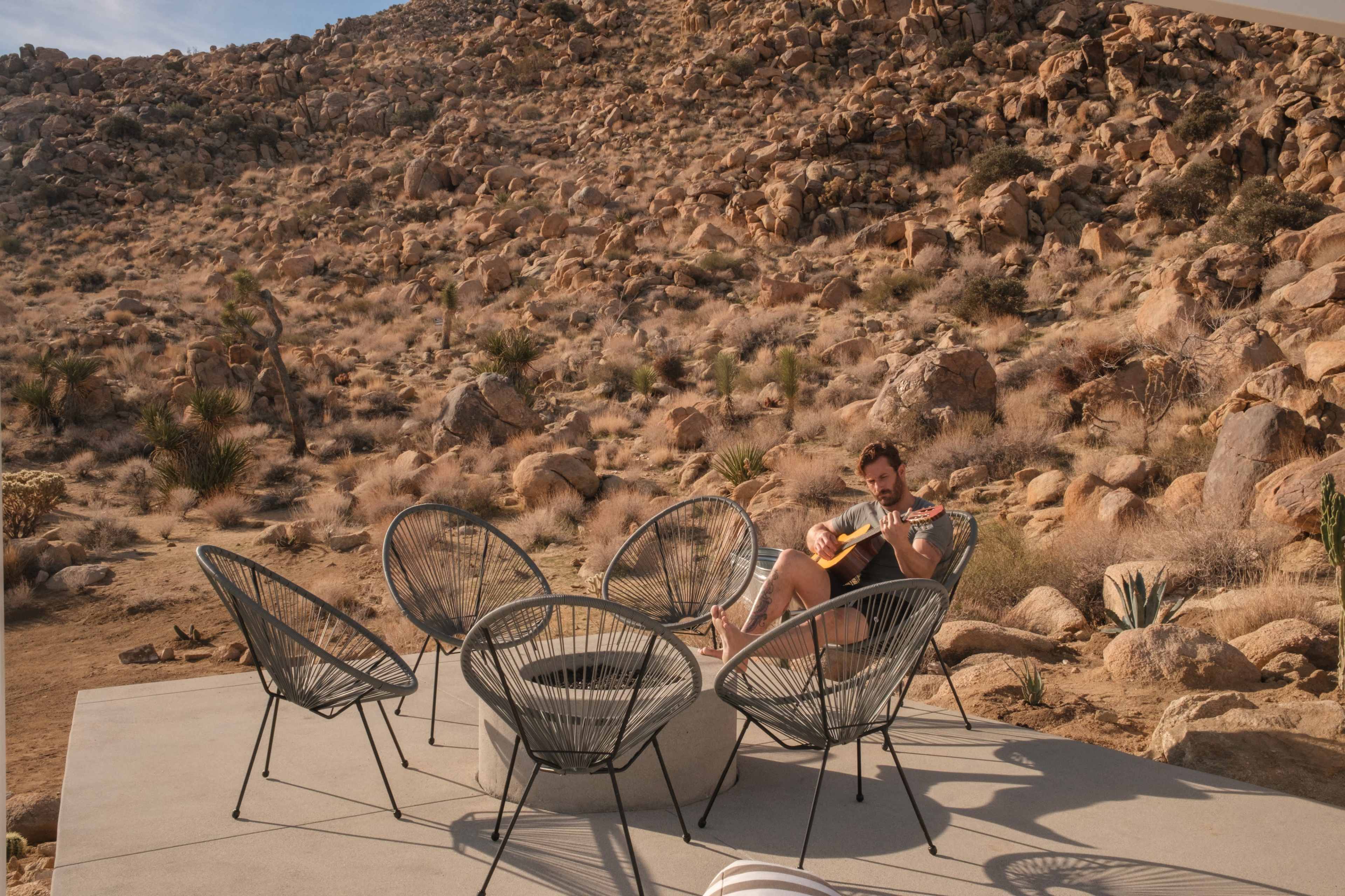 A man sits on a concrete patio playing a guitar surrounded by five chairs in a rocky desert landscape.