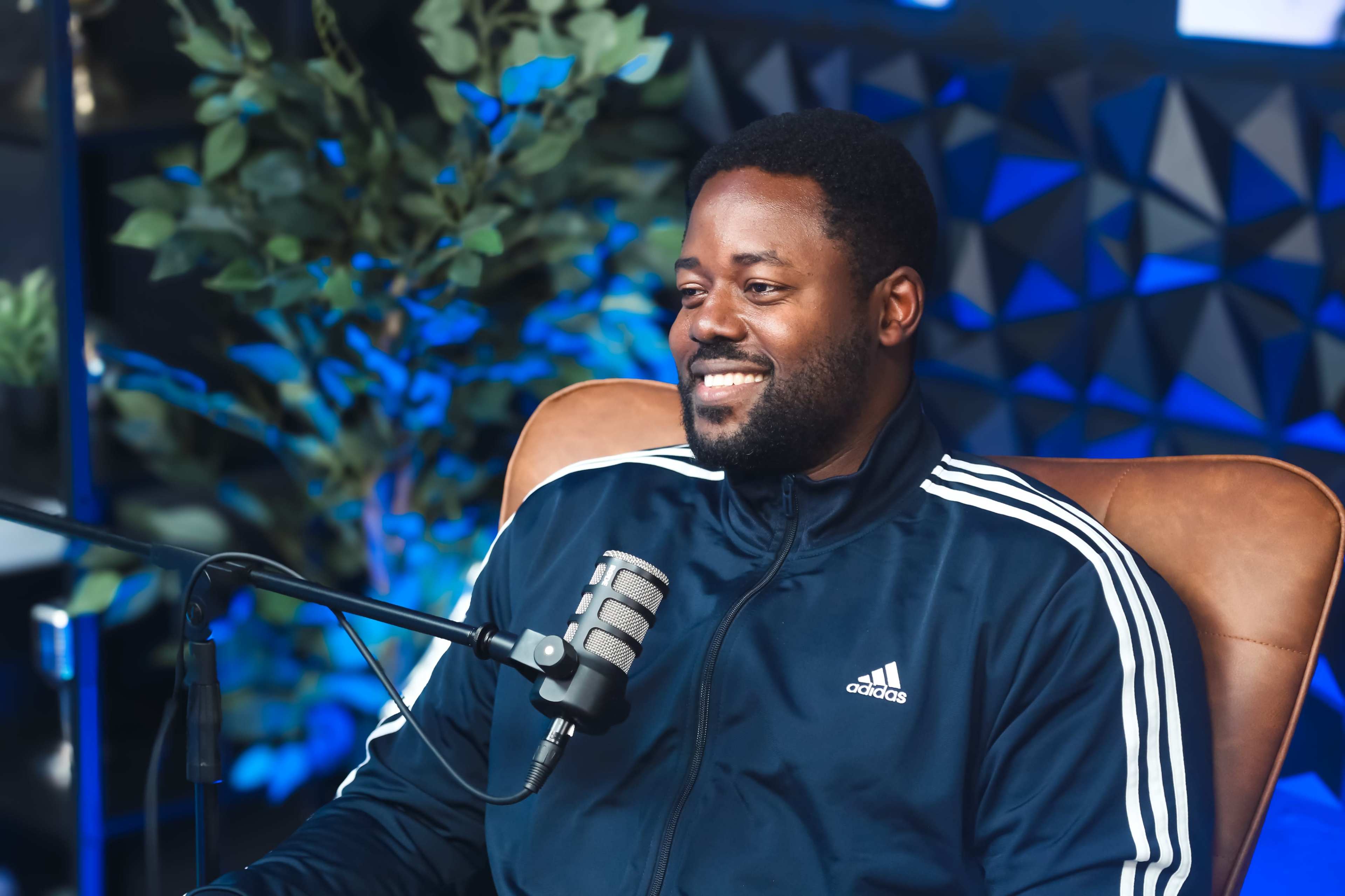 A man in a black and white Adidas tracksuit smiles while sitting in a chair next to a microphone in a modern studio setting.