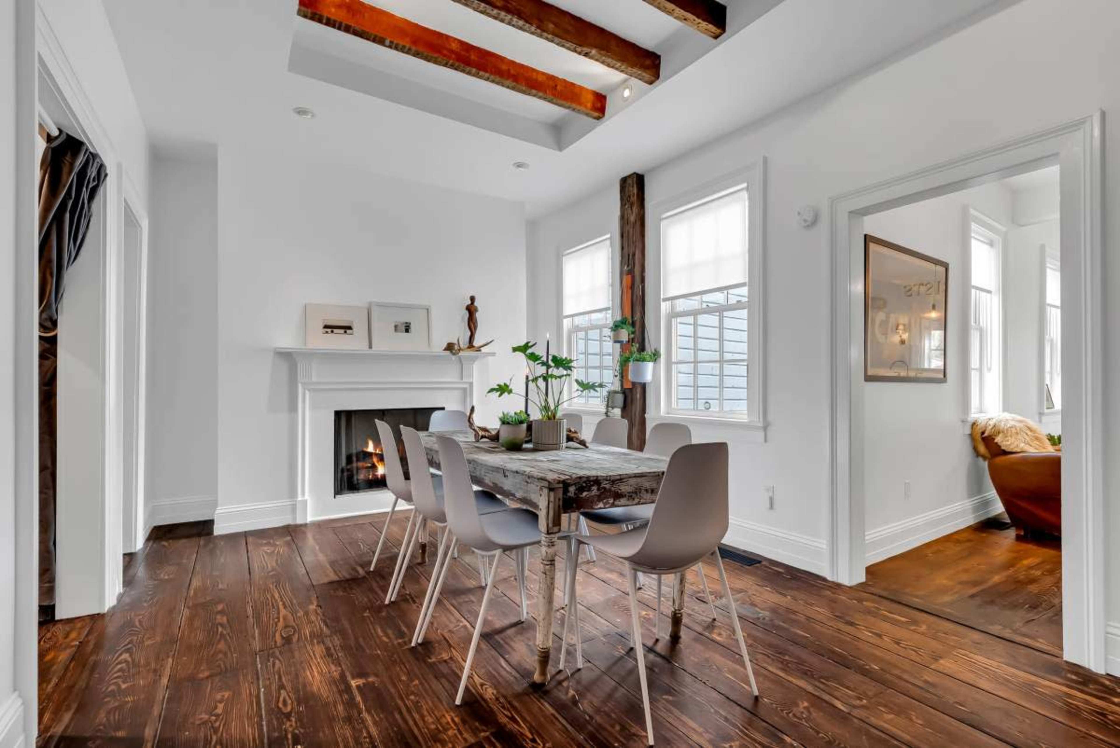 The image shows a minimalist dining area with a wooden table surrounded by white chairs, a fireplace, and exposed wooden beams overhead.