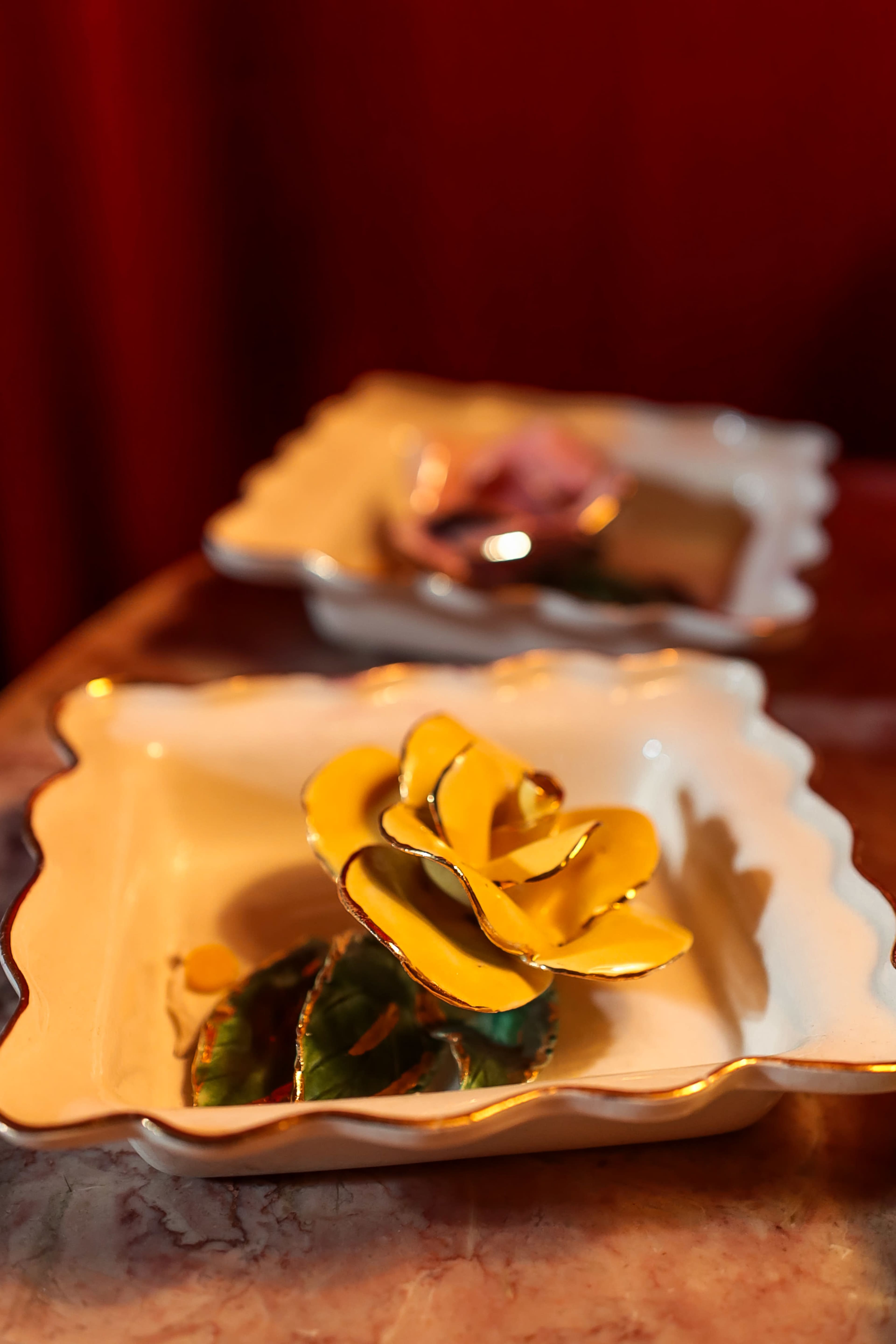 Two decorative ceramic dishes, one featuring a yellow flower and the other a pink flower, rest on a table with a red backdrop.