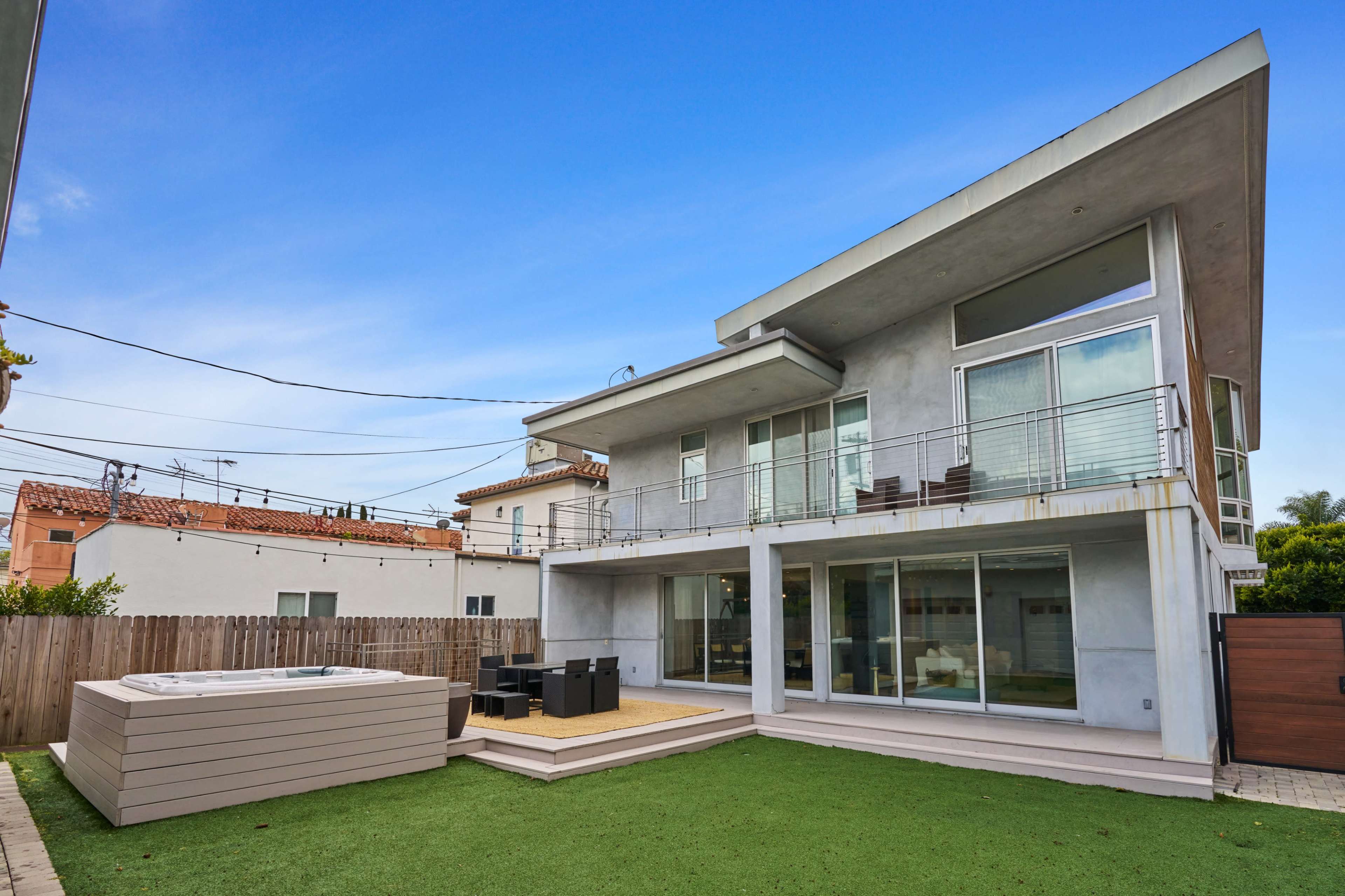 The image shows a modern two-story house with large windows, a minimalist design, and a hot tub on a grassy backyard.