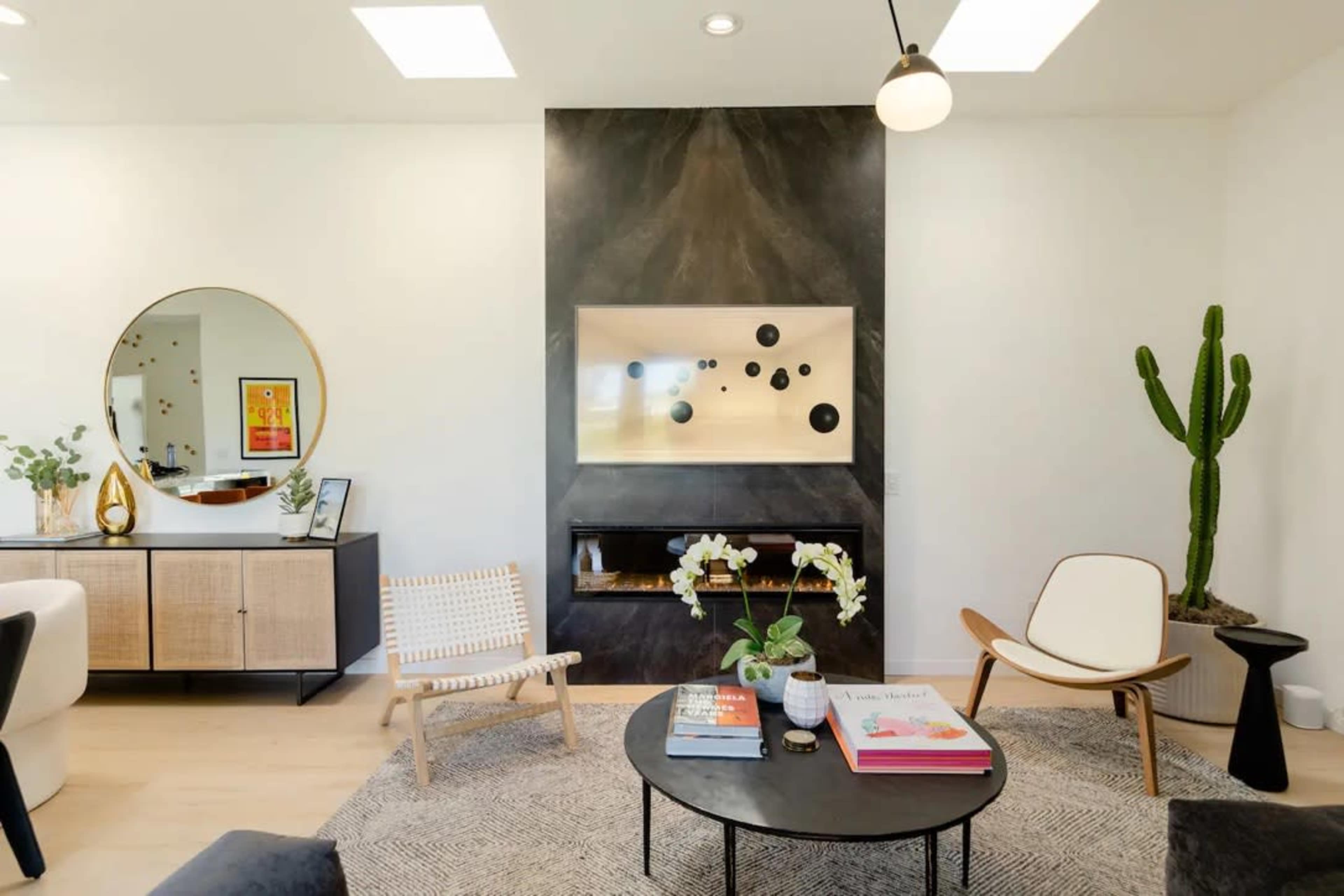 The image shows a modern living room featuring a minimalist fireplace, a round mirror, a cactus plant, and a variety of seating arrangements.
