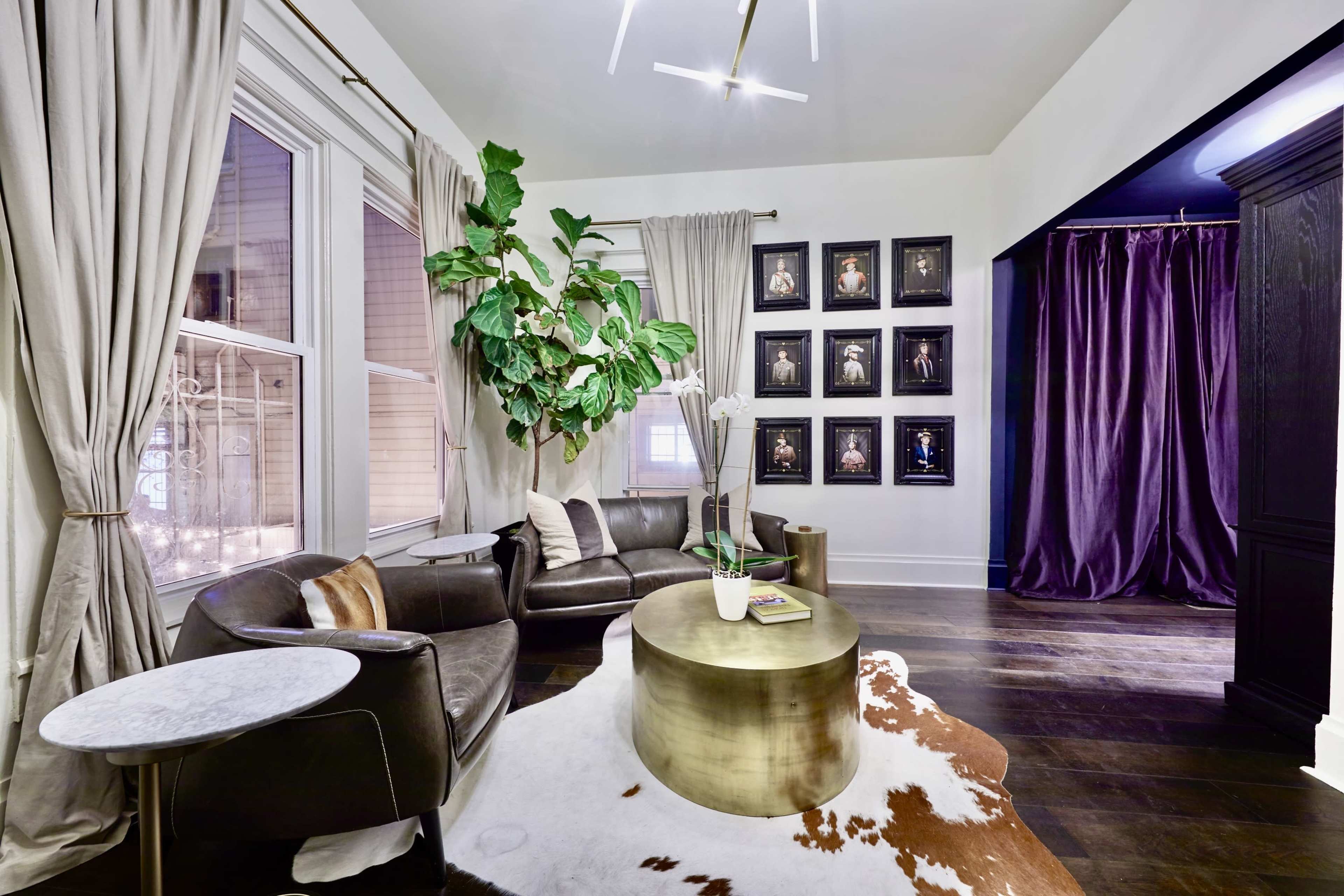 A modern living room features a dark leather sofa, a round metallic coffee table, large plants, and framed artwork on the wall.